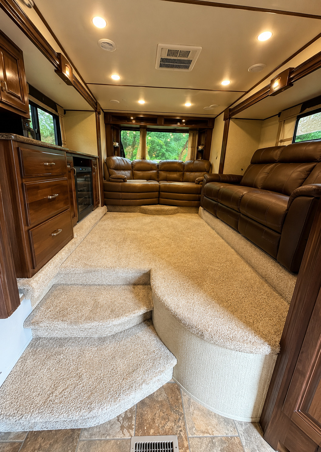 Spacious luxury RV living area featuring brown leather sofas, dark wood cabinetry, carpeted raised flooring and steps, recessed lighting, ceiling vents, large windows, and built-in storage surrounding the comfortable interior.