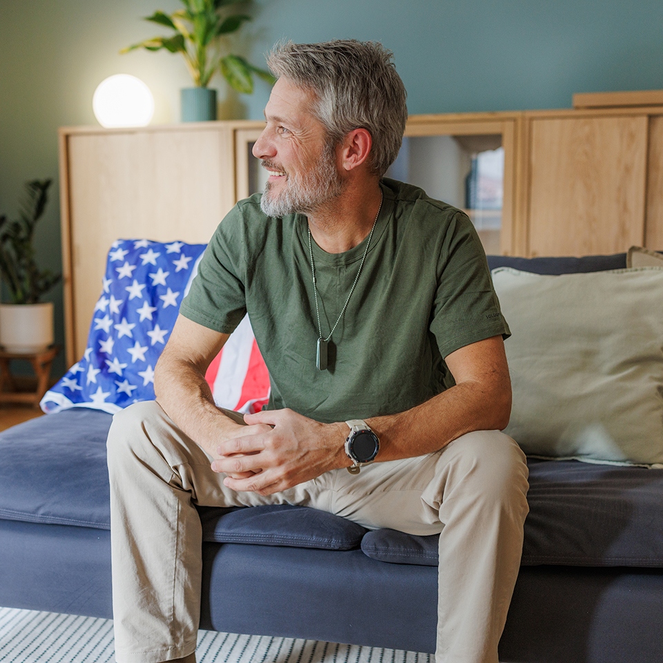 A man wearing a dog tag sitting on a couch with the American flag behind him.