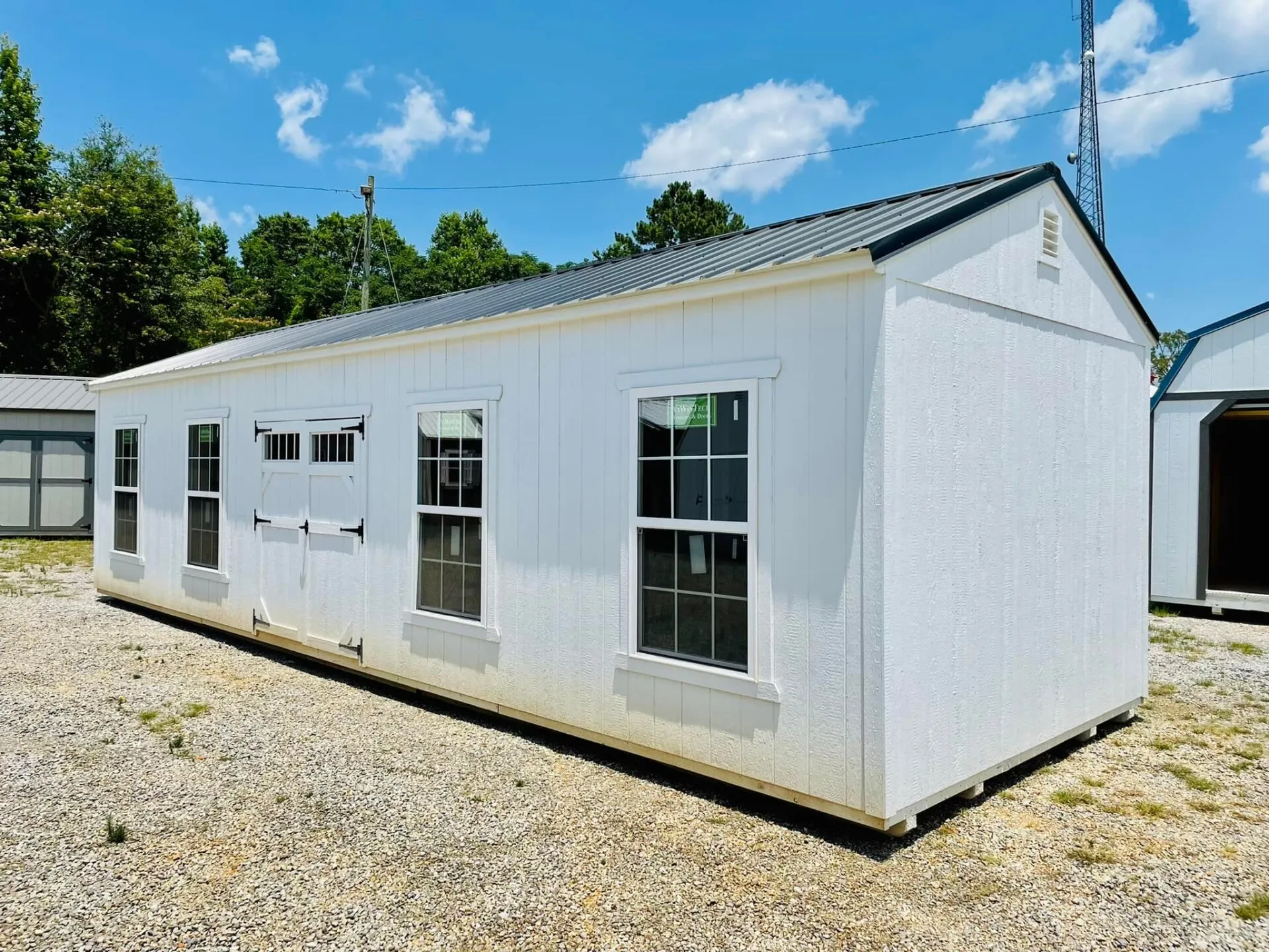 Modern white portable utility building with black metal roof, tall windows, decorative barn-style shutters, and spacious layout. Displayed outdoors, offering attractive storage or workspace functionality for property owners.