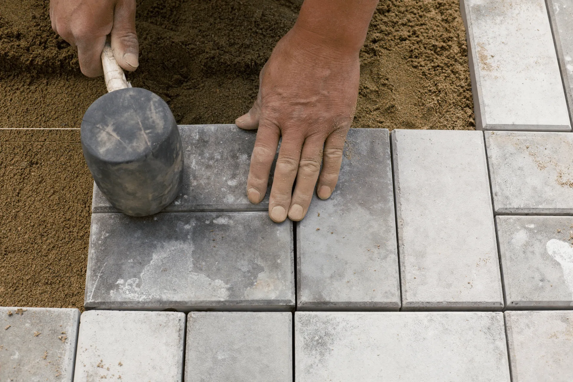 A person's hands are shown using a rubber mallet to adjust paving stones, with sand surrounding the stones.