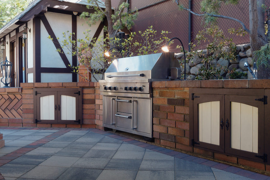 Outdoor patio kitchen with a stainless-steel gas grill, brick counters, wood cabinets, and lush landscaping in a warm, inviting setting.