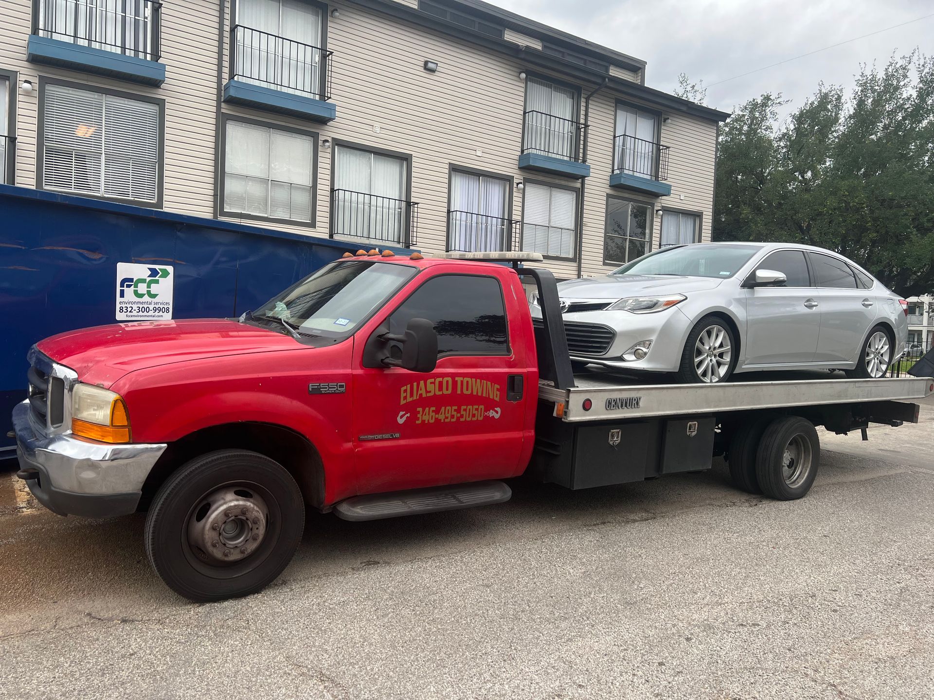 An Eliasco Towing flatbed truck carries a silver Toyota Avalon sedan, parked in front of an apartment complex with balconies and an FCC Environmental Services dumpster nearby.