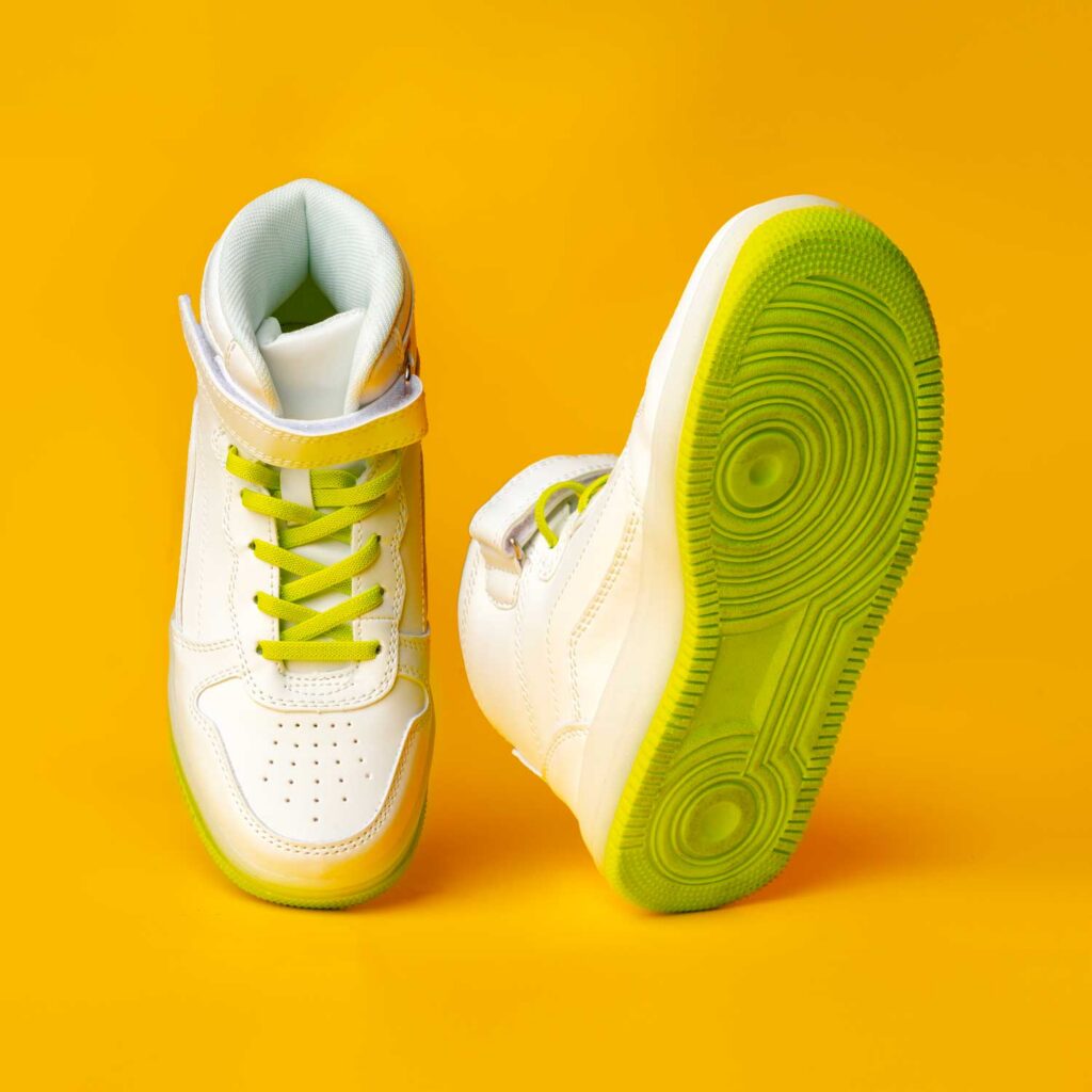 A pair of white high-top sneakers with bright green laces and soles are shown against a vibrant yellow background. The shoes are positioned dynamically, with one shoe facing forward and the other tilted on its side, showcasing its sole.