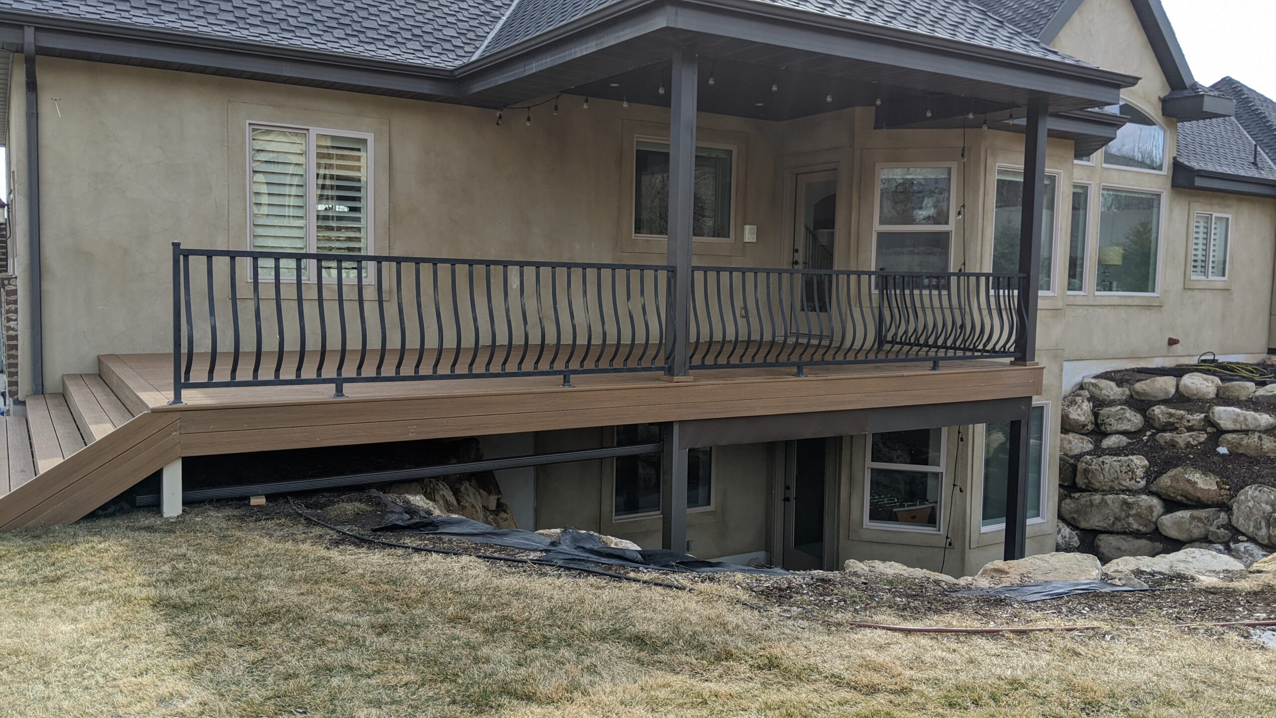 Expand your outdoor living possibilities with this custom deck by DeckPro LLC, featuring low-maintenance composite decking and a highly detailed railing system. The warm brown composite boards provide a durable, splinter-free surface, seamlessly transitioning to wide stairs that connect the home to the large yard. Sleek black metal railings with unique curved balusters wrap around the entire perimeter, offering a modern touch that contrasts beautifully with the home&rsquo;s stucco exterior and brick accents. Pictured on a sunny day, this design illustrates how DeckPro can create a functional, stylish, and safe retreat for any home, regardless of terrain, thanks to a robust structural support system and expertly integrated rock retaining walls.