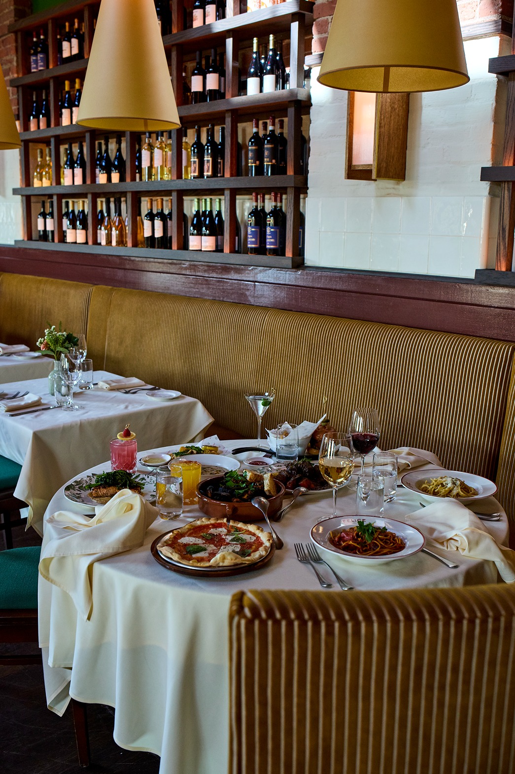 A spread of Italian dishes, including pasta and appetizers, sits on a table by a large window. The bright, airy space features cafe curtains and elegant glassware, creating an inviting lunch atmosphere.