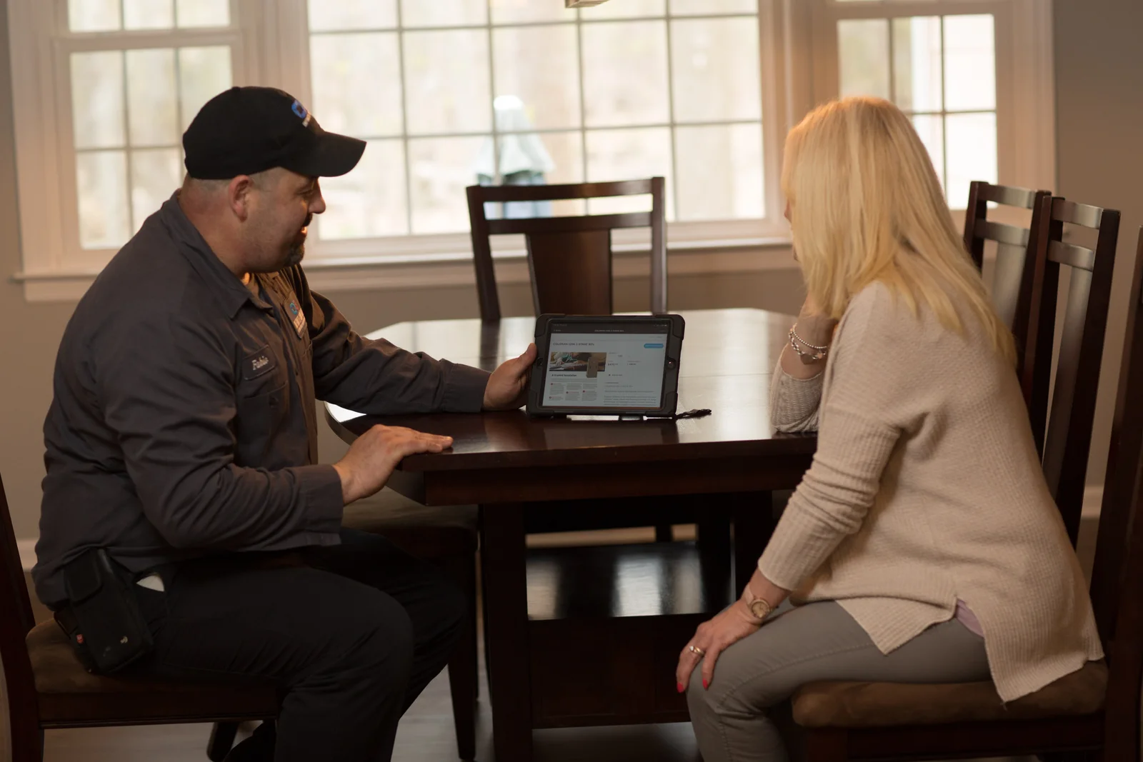 A man in a dark grey uniform and a black baseball cap is showing something on a tablet to a blonde woman sitting across from him at a dark wooden dining table. The tablet screen displays a website with product listings and images. The man has his left hand on the table and is holding the tablet with his right. The woman is resting her chin on her left hand, looking at the tablet. The scene is indoors, with large windows behind them showing natural light and trees outside.