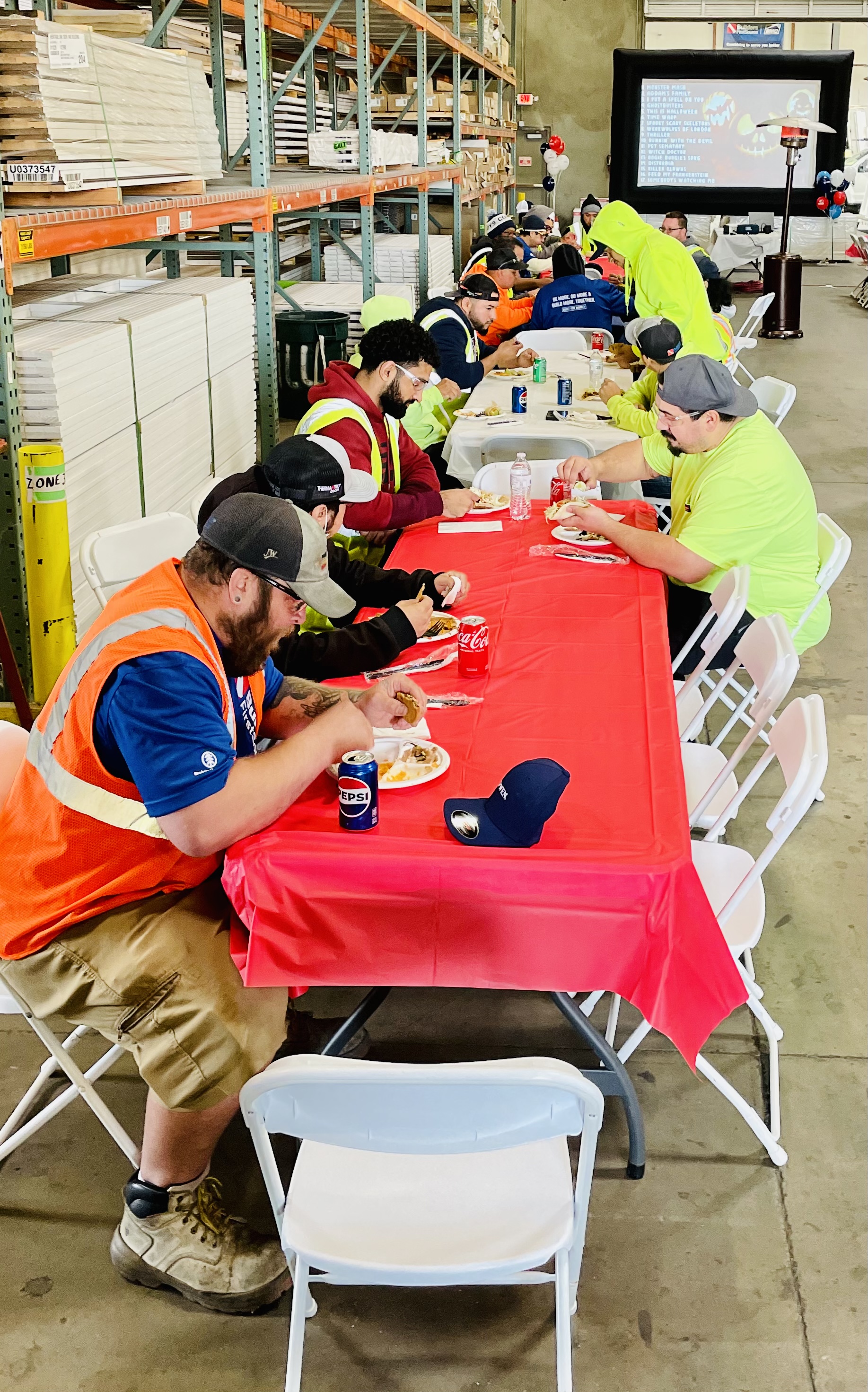 A group of people wearing safety vests and casual clothing are sitting at long tables covered with red tablecloths, eating and drinking. The tables are set up in a warehouse with stacks of materials and a large screen in the background.