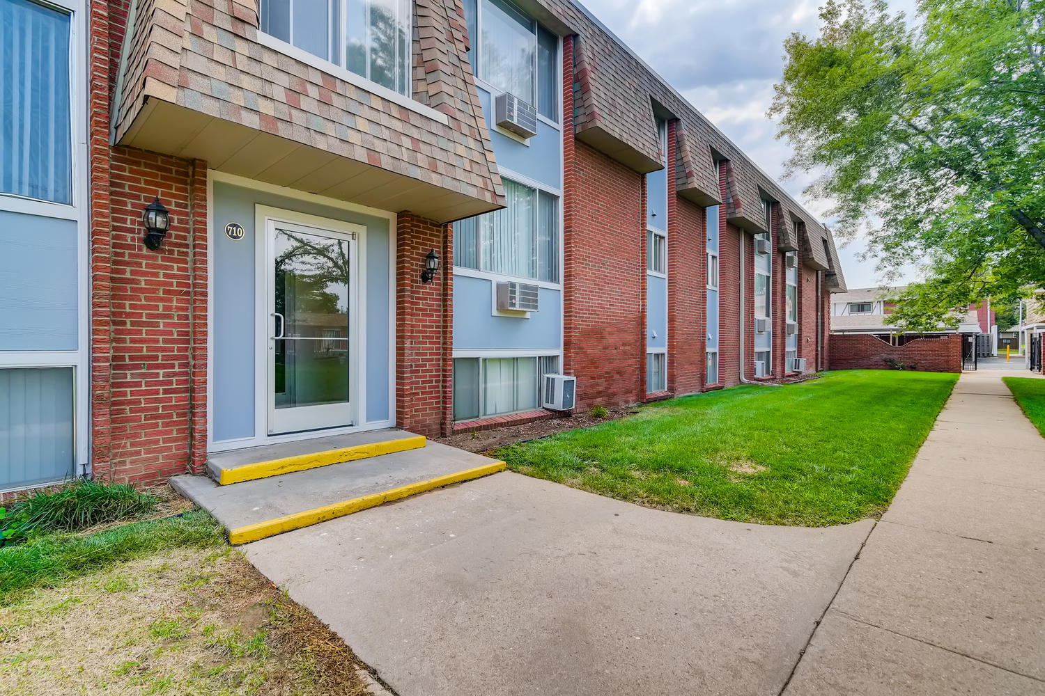 Glass door building entrance with brick and siding building exterior and grass lawn with sidewalk.