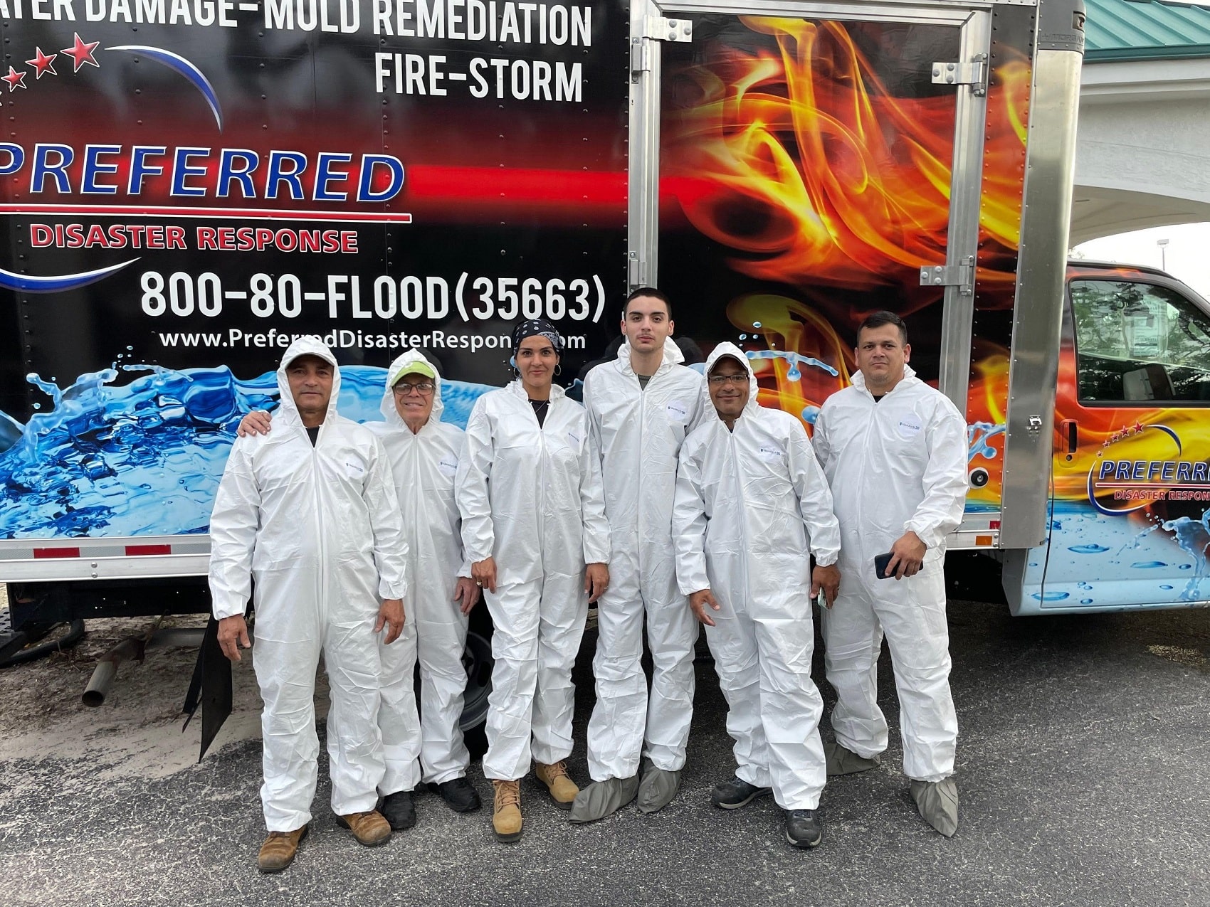 A team of six people wearing white protective suits stands in front of a disaster response truck. The truck has graphics of water and fire on its sides and displays the company name "Preferred Disaster Response" along with a phone number. The team members are of diverse backgrounds and ages, with some smiling and others looking directly at the camera. They are standing on a paved surface, possibly a parking lot. The sky appears overcast.