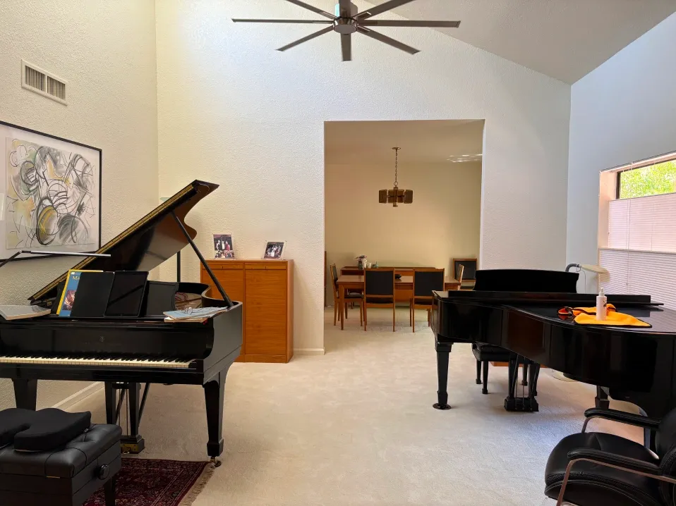 Elegant living room with vaulted ceiling, featuring two grand pianos, soft carpeting, modern ceiling fan, and open view into dining area, creating a refined, spacious, and music-focused interior environment.