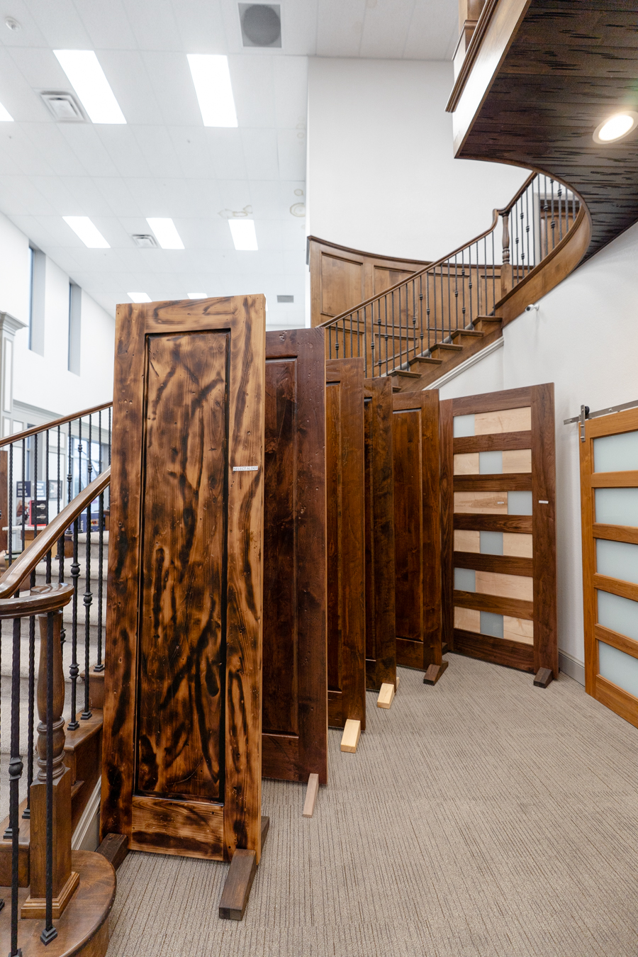 Wooden doors displayed in a Builders FirstSource showroom, with a curved staircase in the background.