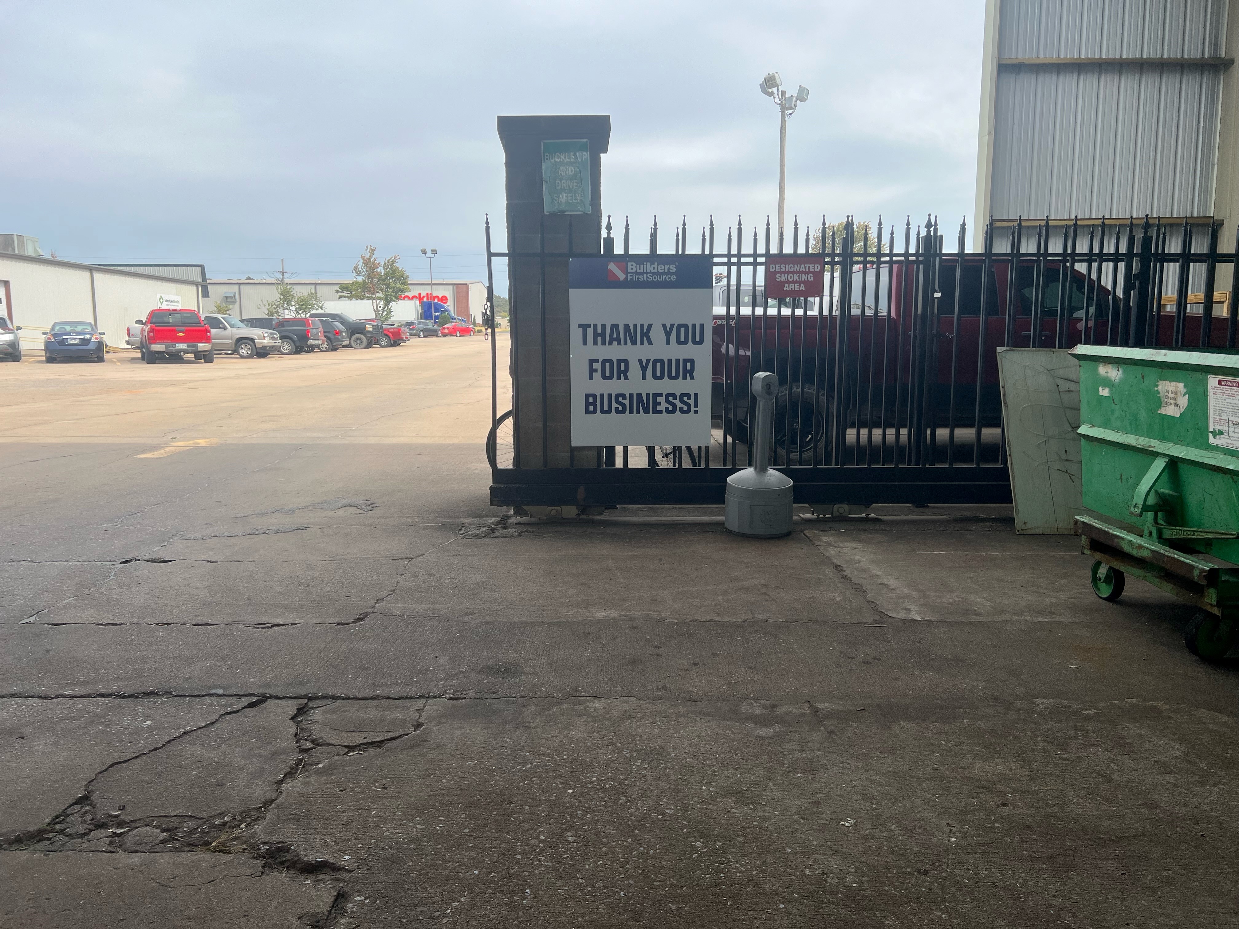 A gated exit at a Builders FirstSource facility with a sign that reads "Thank you for your business!" The gate is black with metal bars, and a "Designated Smoking Area" sign is visible nearby. In the background, a parking lot with several vehicles, including a red truck and a green dumpster, can be seen. The sky is overcast, adding a muted tone to the scene.