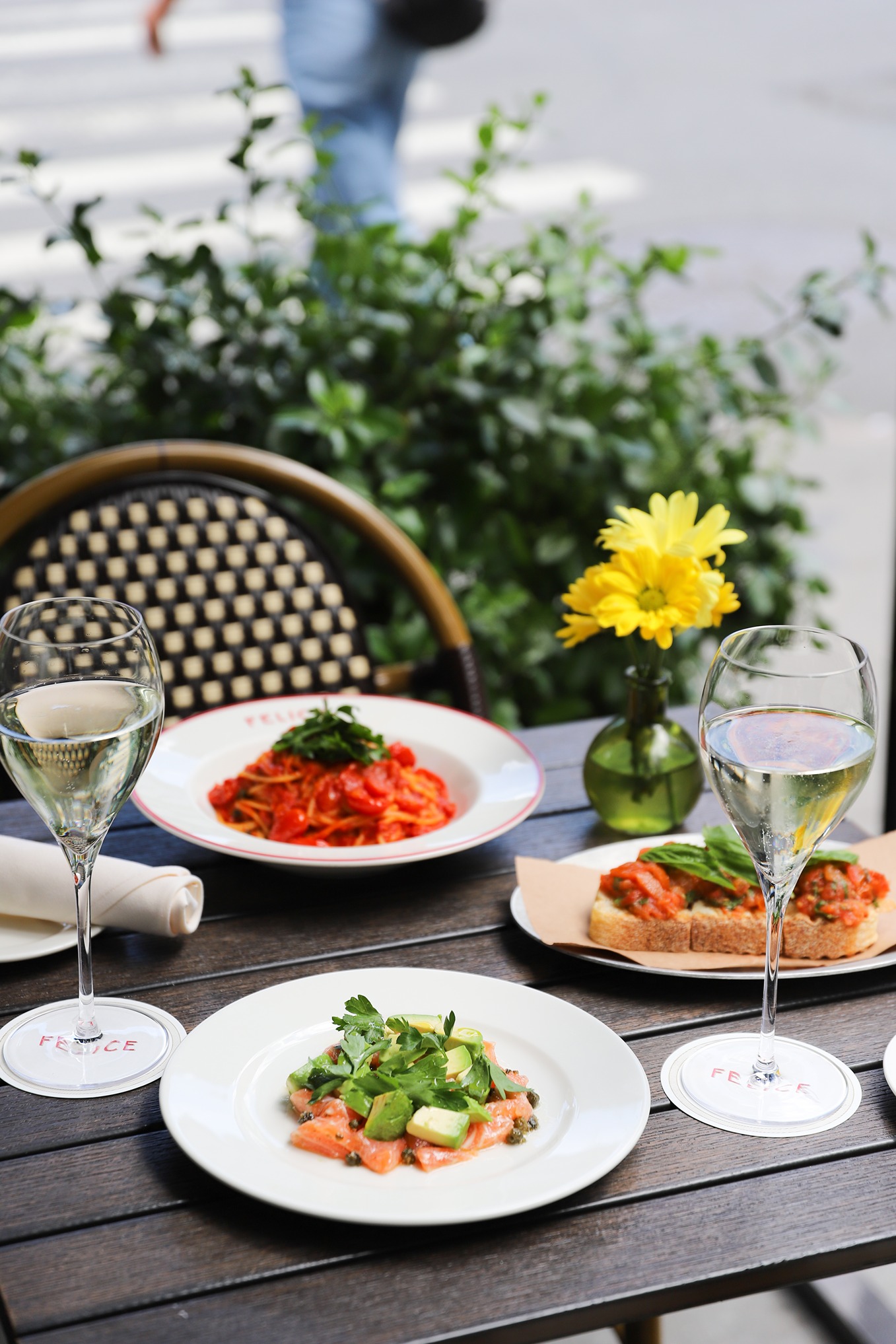 An al fresco setting featuring spaghetti pomodoro, salmon tartare with avocado, and bruschetta. Two glasses of white wine sit on branded Felice coasters on a dark wooden table.