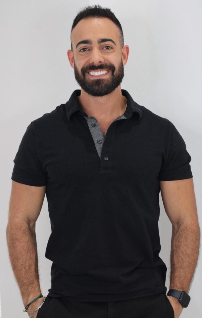 A man with a beard smiles at the camera, wearing a black polo shirt with a gray collar and a smartwatch on his left wrist. His hands are in his pockets.