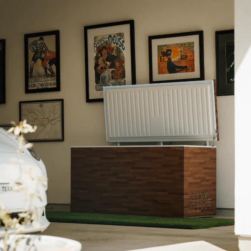 An open chest freezer with a wood-grain exterior is situated on a patch of artificial grass in what appears to be a garage or workshop. The freezer's lid is open, revealing a white ribbed interior. Several framed vintage posters and a map are mounted on the wall behind the freezer. A white car is partially visible in the foreground on the left side of the image.