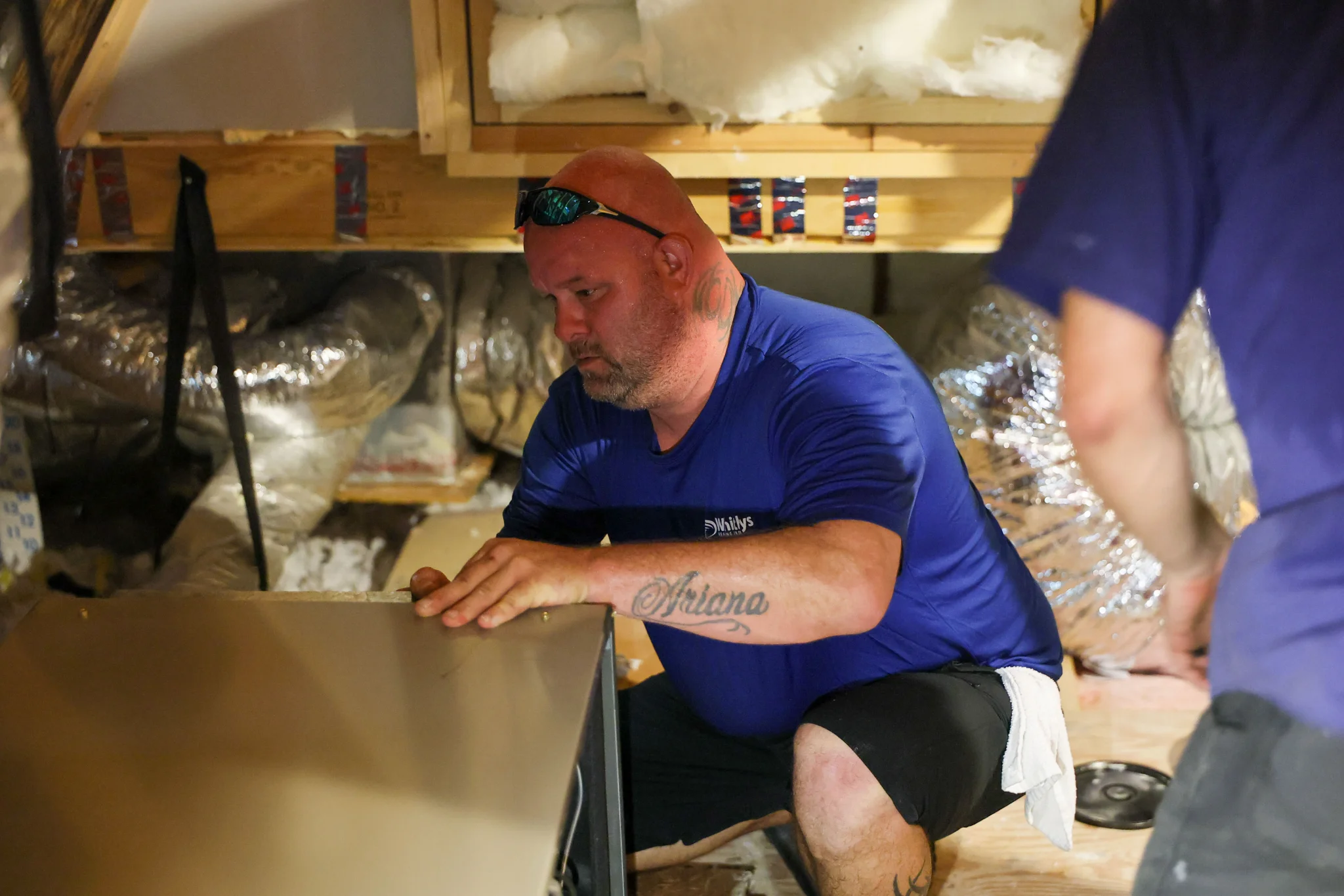 A man with a shaved head and sunglasses on his head is working in what appears to be an attic. He is 