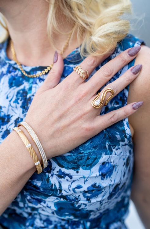 Gold-toned leaf and curved statement rings pair with sparkling bracelets and a chain necklace against a cobalt floral dress. Explore curated jewelry at Eliana Curated in Old Town Alexandria, Virginia.