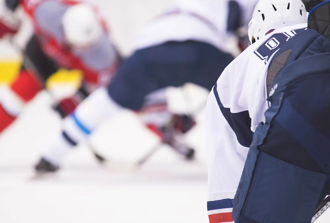 A hockey game scene showing a player in a white jersey in the foreground, with three players clashing in blurred motion behind, conveying dynamic action.