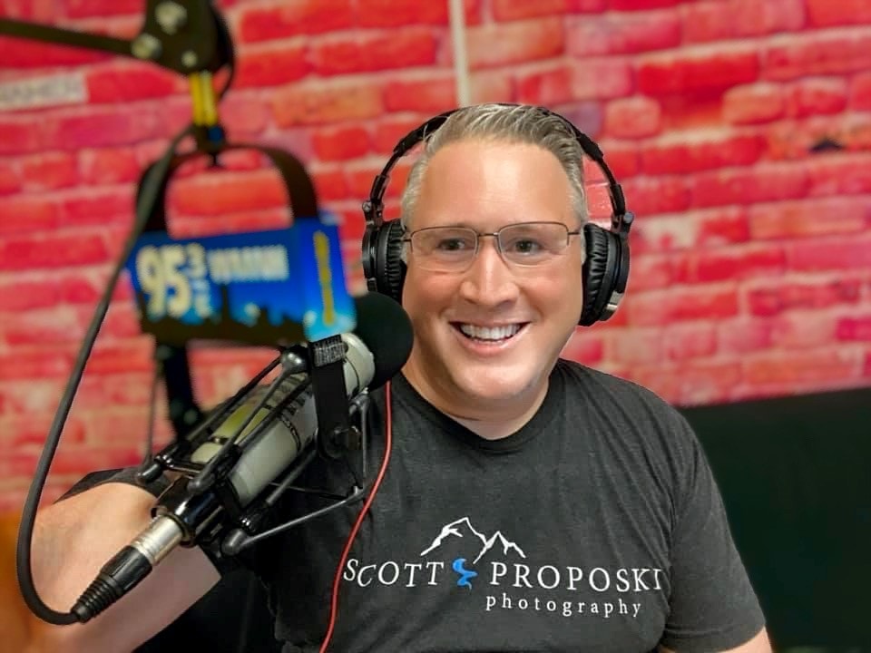 A smiling man with glasses and headphones on is sitting in front of a microphone in a recording studio. He is wearing a dark t-shirt with white text and graphics that read 'SCOTT PROPOSKI photography' and a mountain logo. In the background, a brick wall is visible, and a blue sign with the numbers '95.3' and a city skyline graphic is attached to a microphone stand.