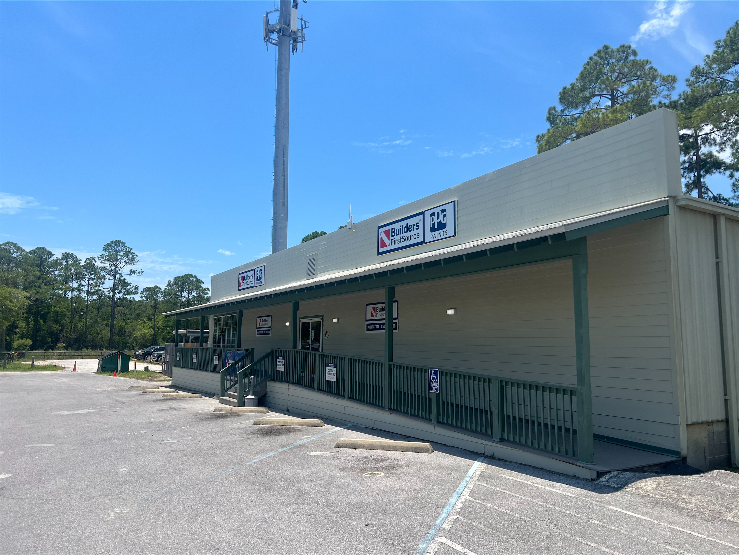 The image depicts the exterior section of the "Builders FirstSource" hardware store. This part of the building is also a single-story structure with a green and beige color scheme. The "Builders FirstSource" sign is prominently displayed on the upper front wall, next to a sign for "PPG Paints."
There is a ramp leading up to the entrance, which is accessible and has handrails. The parking area in front of the building includes designated handicap parking spaces. The surroundings feature a large parking lot and tall pine trees in the background. Additionally, a tall cell tower is visible behind the building. The sky is clear and blue, indicating a sunny day.