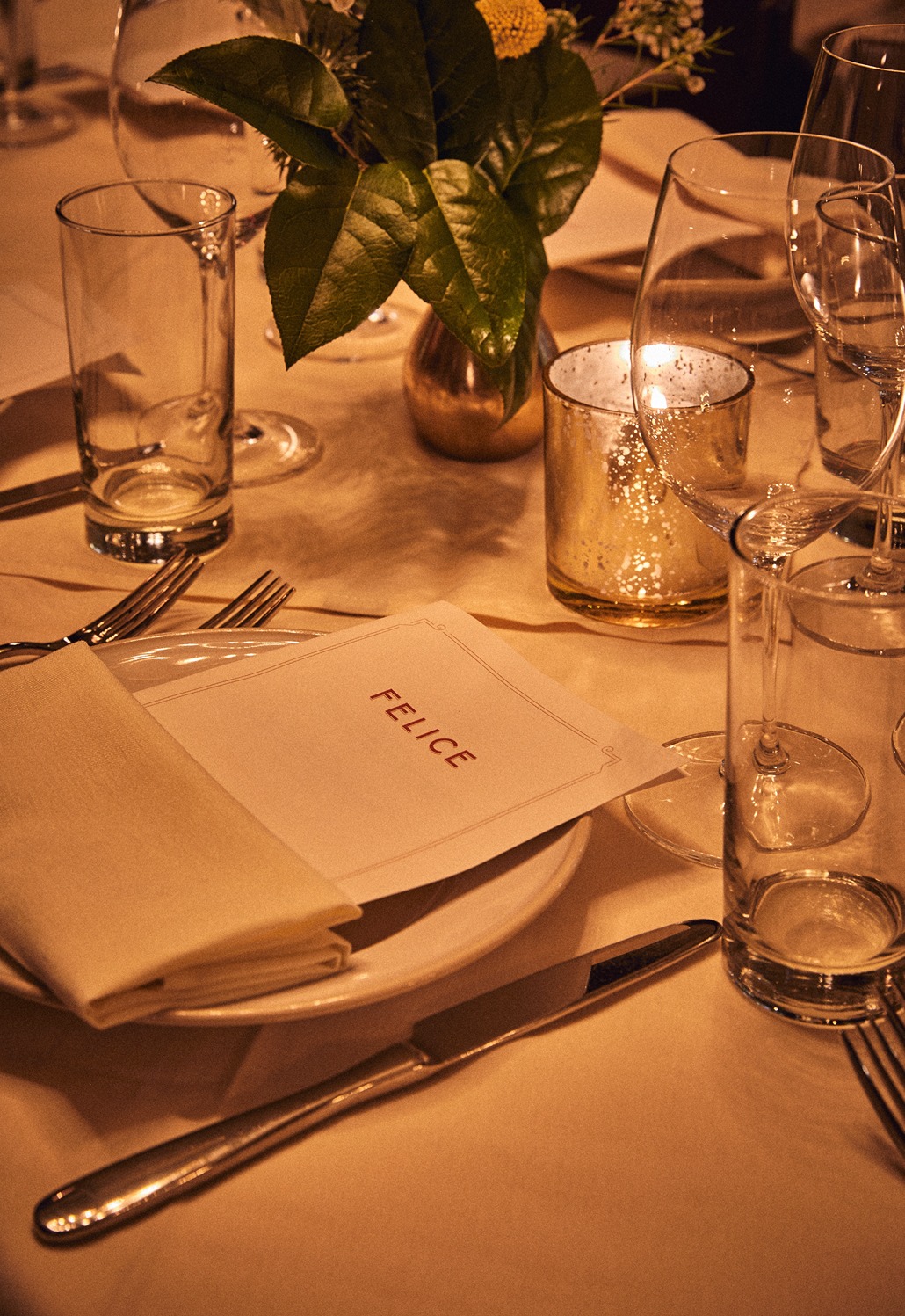 A close-up of a sophisticated table setting featuring a white plate, a folded napkin, and a menu with the "FELICE" logo. A flickering mercury glass candle and a small floral arrangement create a warm, golden glow across the glassware.