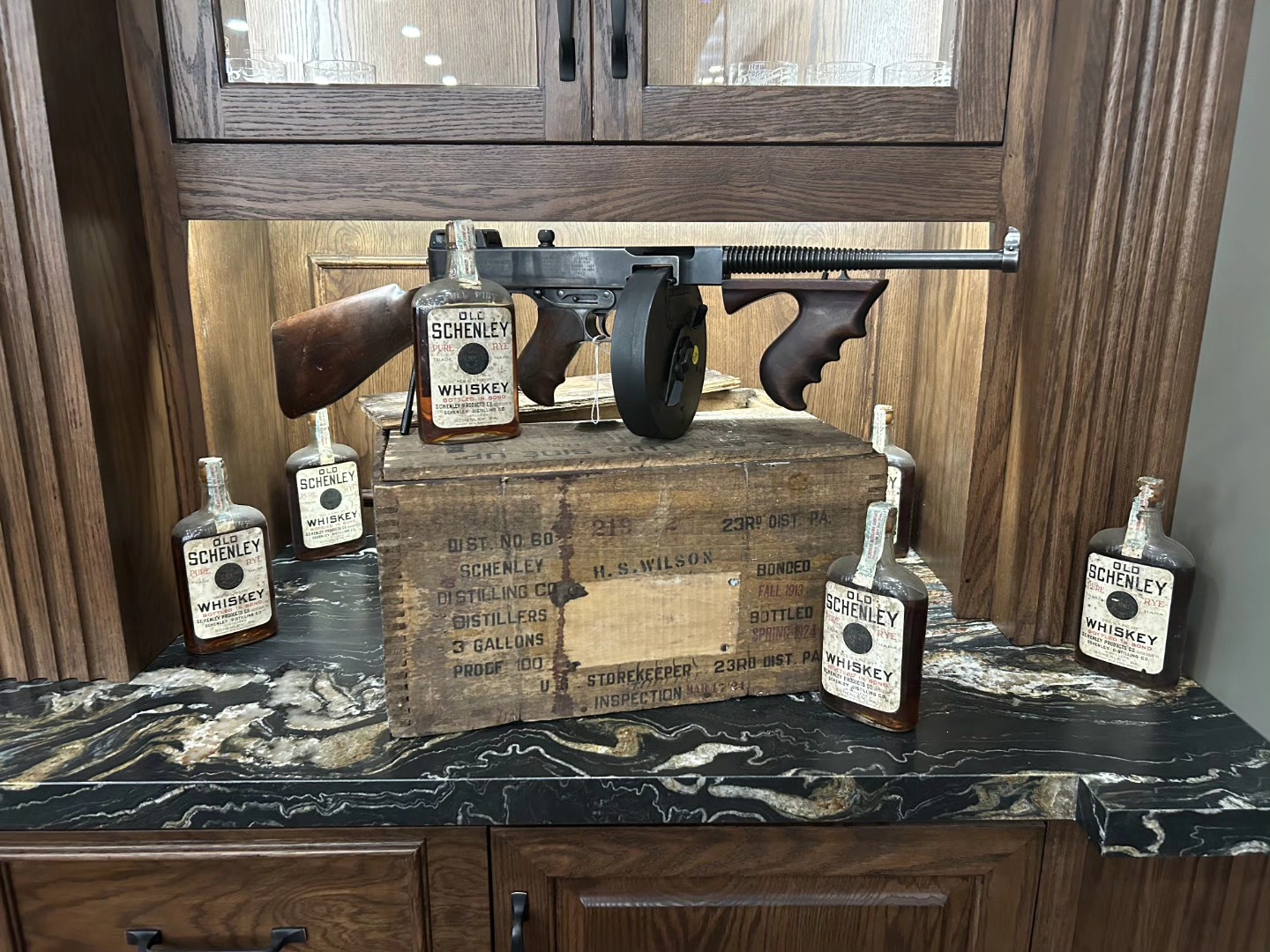 A historic Thompson submachine gun presented alongside Prohibition-era Old Schenley whiskey bottles and a period wooden crate — a signature display piece at Shoot It LLC's Phoenix showroom.