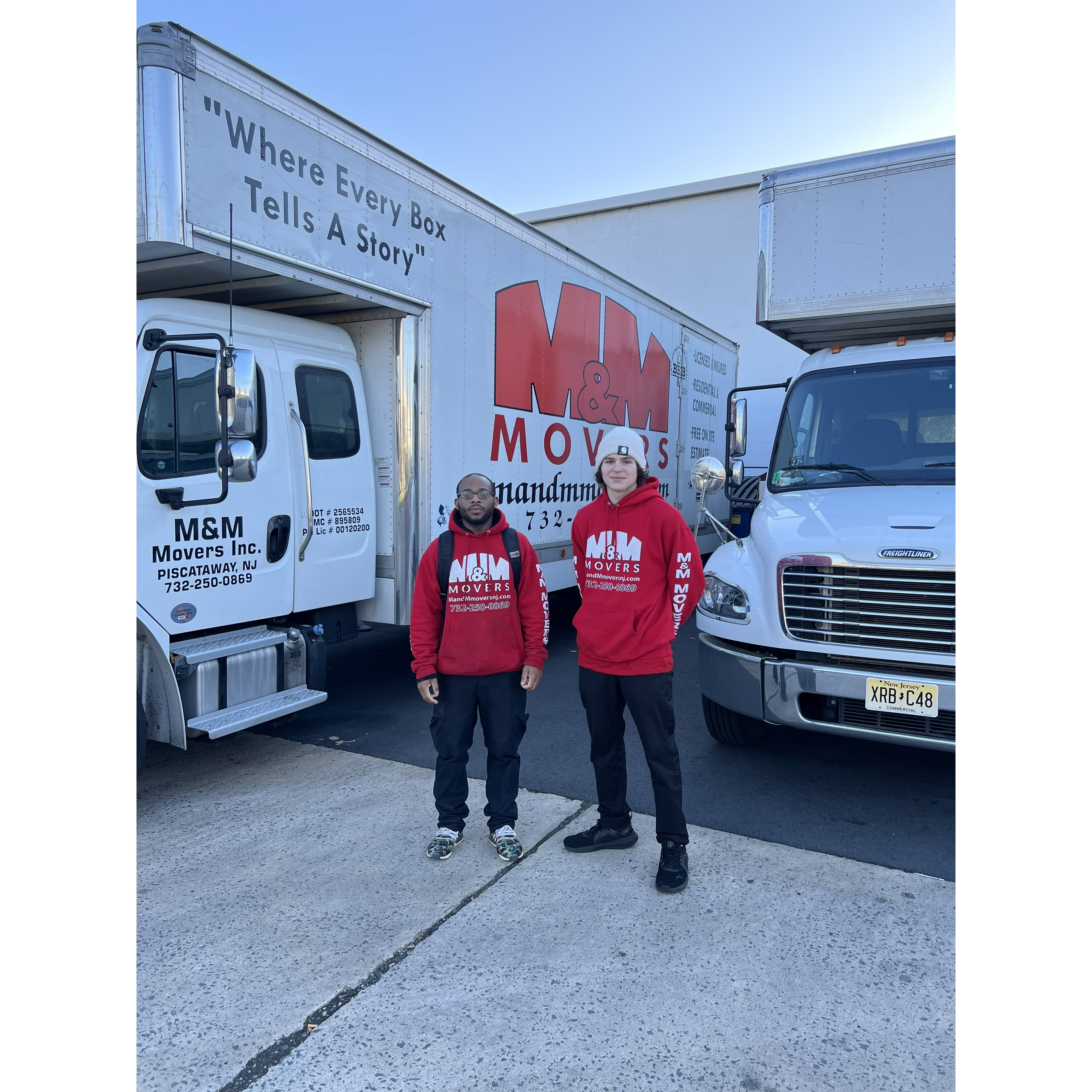 Two movers in matching red hoodies and black pants stand between two white box trucks—one side bearing the slogan “Where Every Box Tells A Story”—gearing up for the day’s haul.