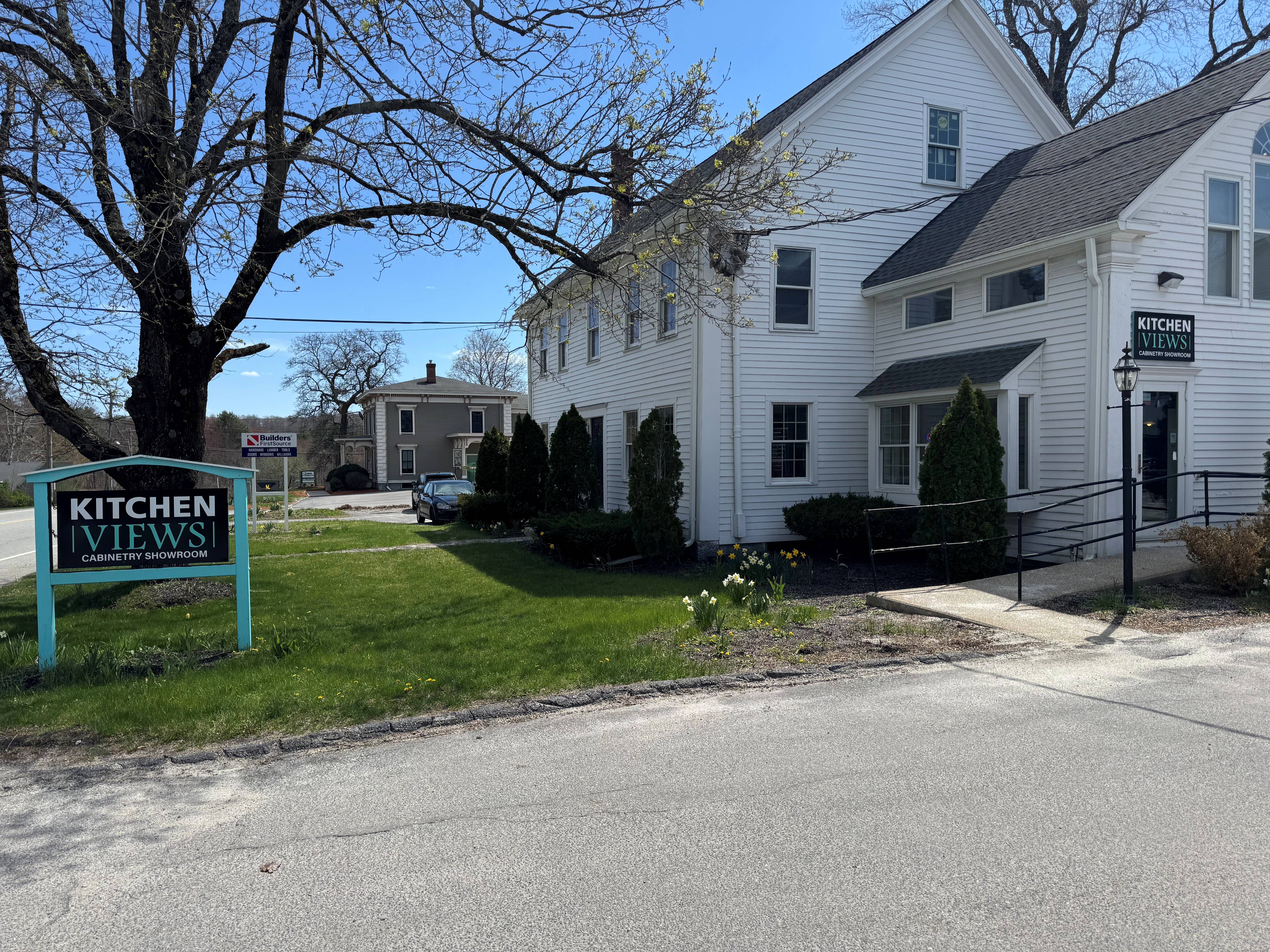 White two-story showroom building with a &lsquo;Kitchen Views&rsquo; sign on a grassy corner, featuring large windows, trimmed shrubs, and a sidewalk leading to the entrance.&rdquo;