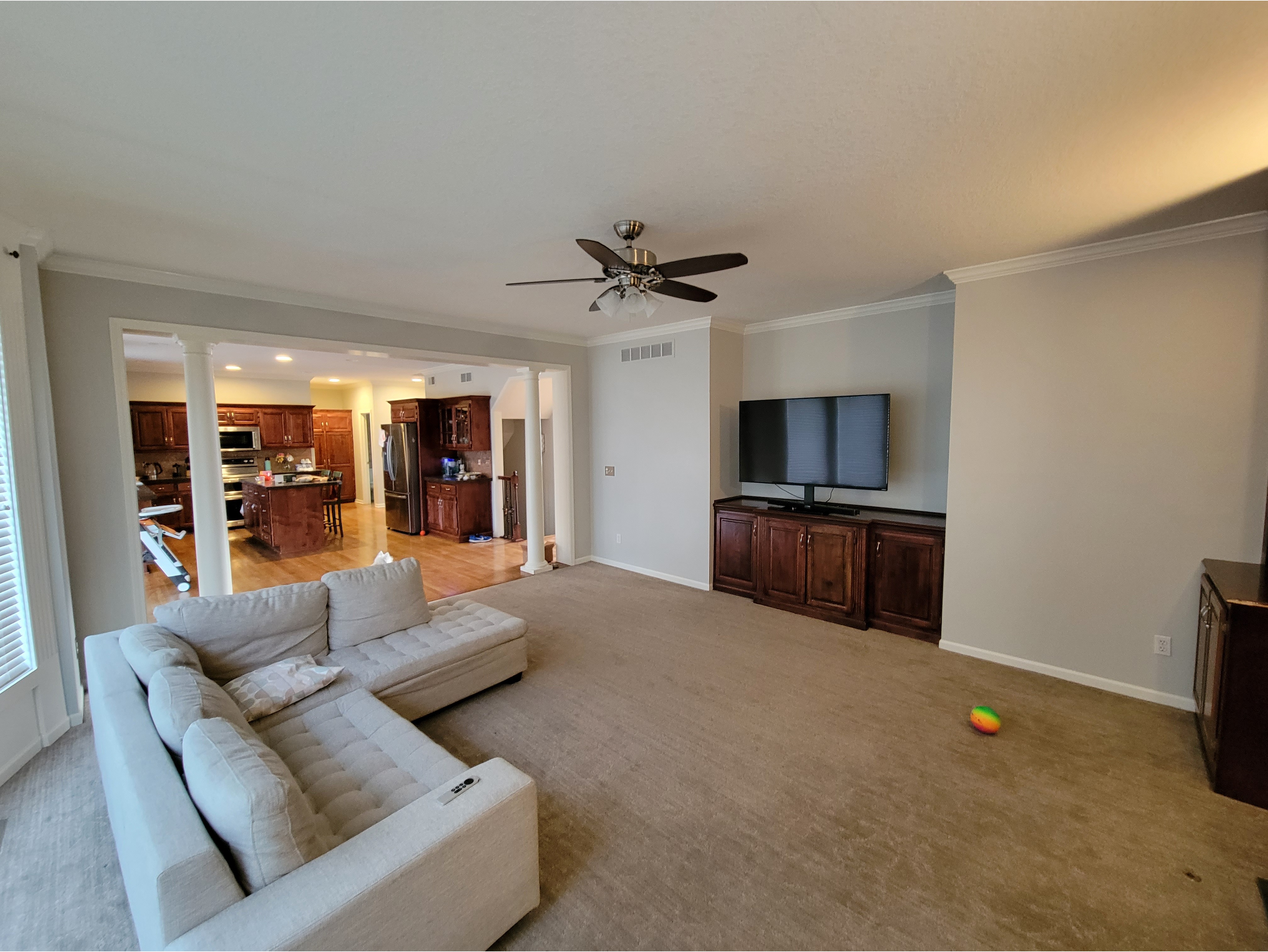 A spacious living room featuring a fresh interior paint job in neutral tones, complemented by crown molding and trim work for a clean and modern look. The open-concept design connects to a kitchen with wooden cabinets and hardwood flooring, creating a seamless flow. Ideal for showcasing interior painting services, trim installation, and open-layout designs.