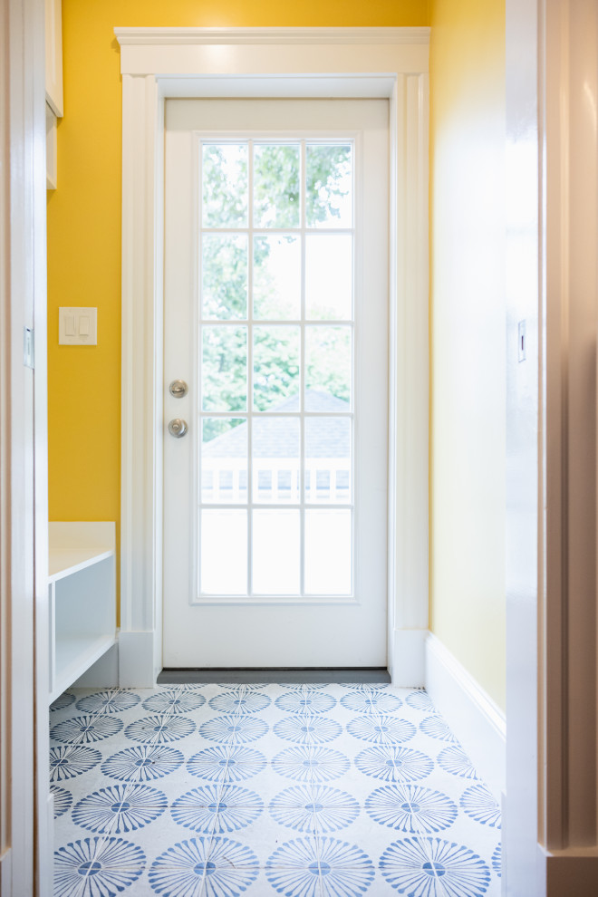 A hallway with bright yellow walls leads to a white paneled glass door. The floor is tiled with a repeating pattern of blue starbursts on a white background. A white bench with shelves is visible on the left side of the hallway.