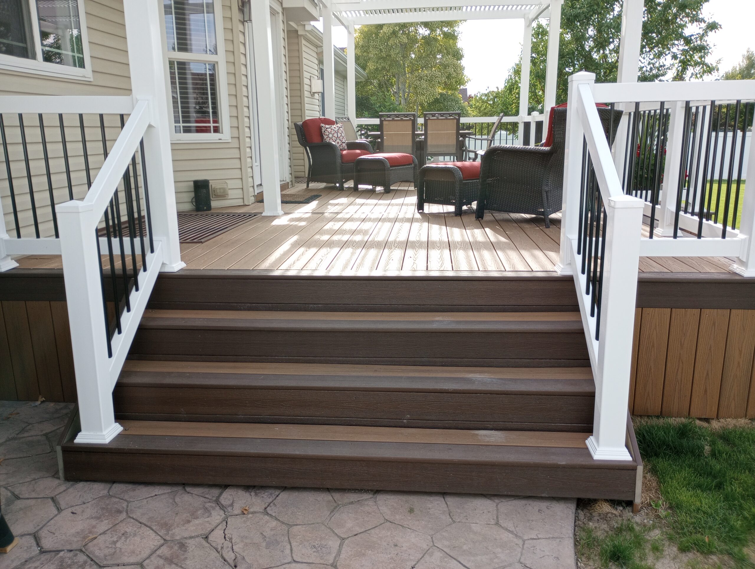 Create the perfect low-maintenance backyard retreat with a custom composite deck from DeckPro LLC. This beautiful project features rich brown composite decking paired with elegant white vinyl railings and contrasting black metal balusters. The attached white pergola provides comfortable shade, while the wide stairs offer an inviting transition to the stamped concrete patio below.