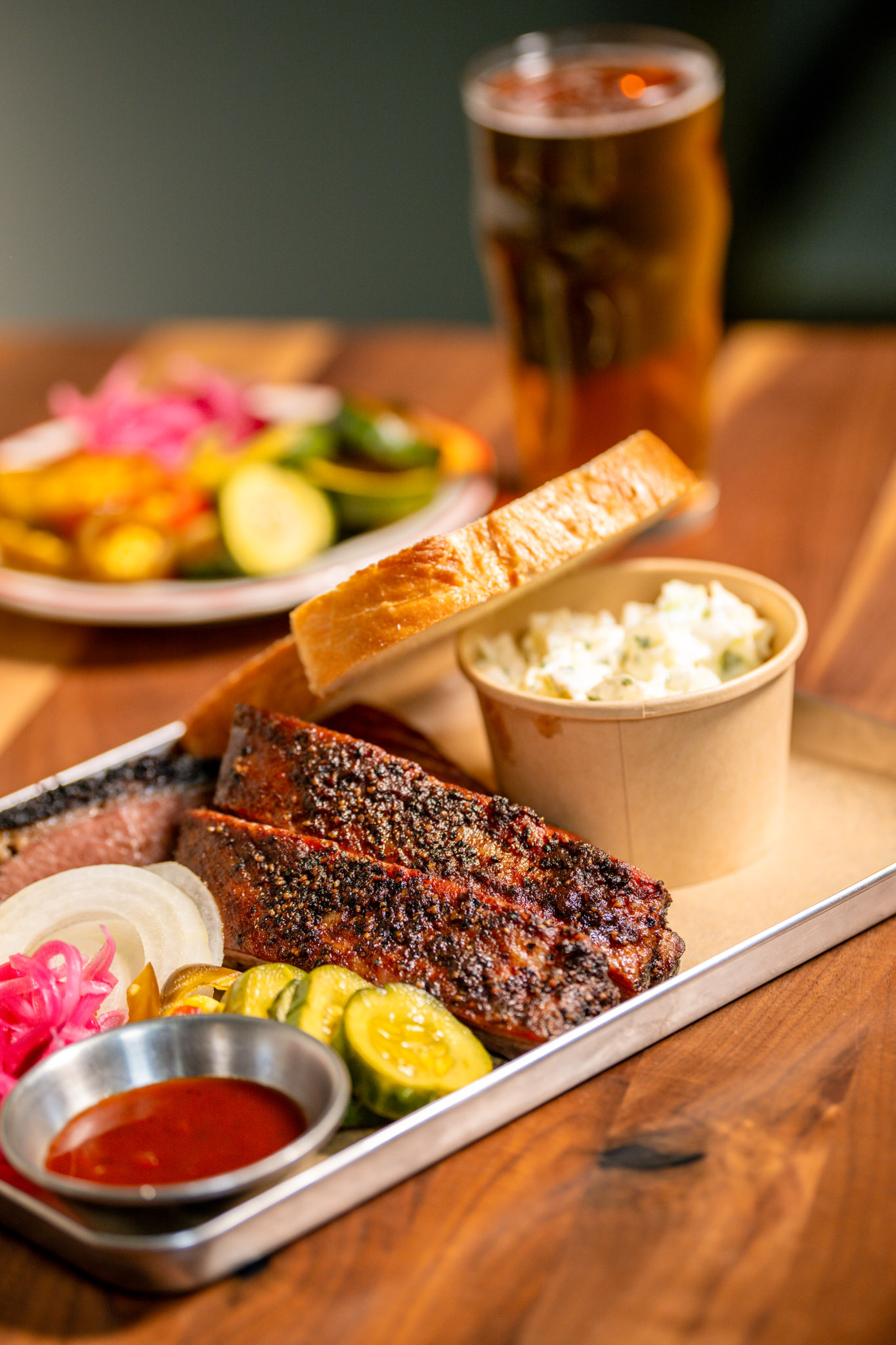 A metal BBQ tray with Sweet Heat St. Louis Spareribs, white bread, coleslaw, pickled vegetables, and Joe's BBQ sauce, served alongside a plate of colorful pickled vegetables and a cold beer in the background.