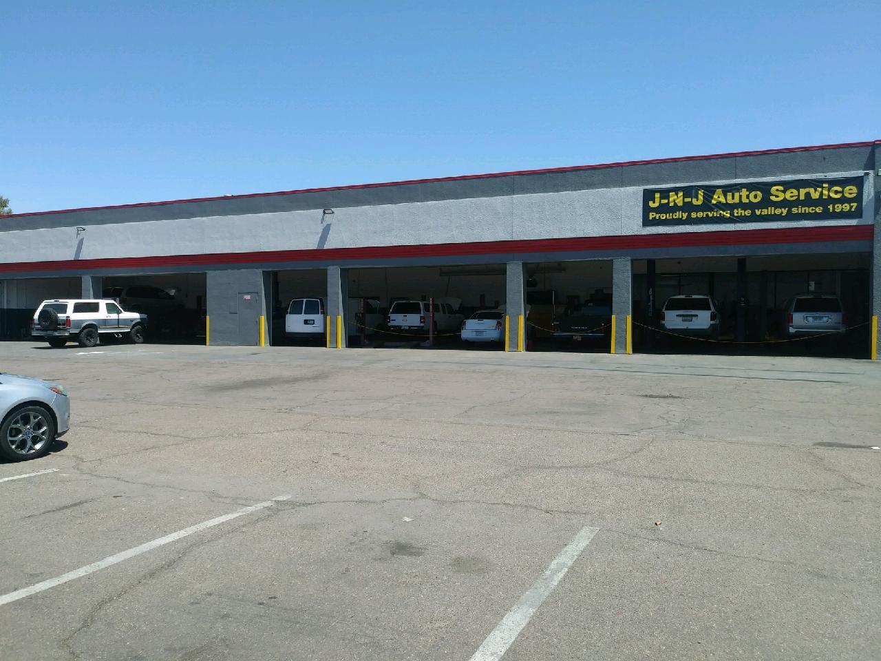 A wide shot of a commercial building with multiple bays for vehicle service. Several vehicles, including vans and SUVs, are parked inside and outside the bays. A sign above the bays reads "J-N-J Auto Service" and "Proudly serving the valley since 1997." The building is gray with red trim, and the sky is clear blue. A parking lot with white lines is in the foreground.