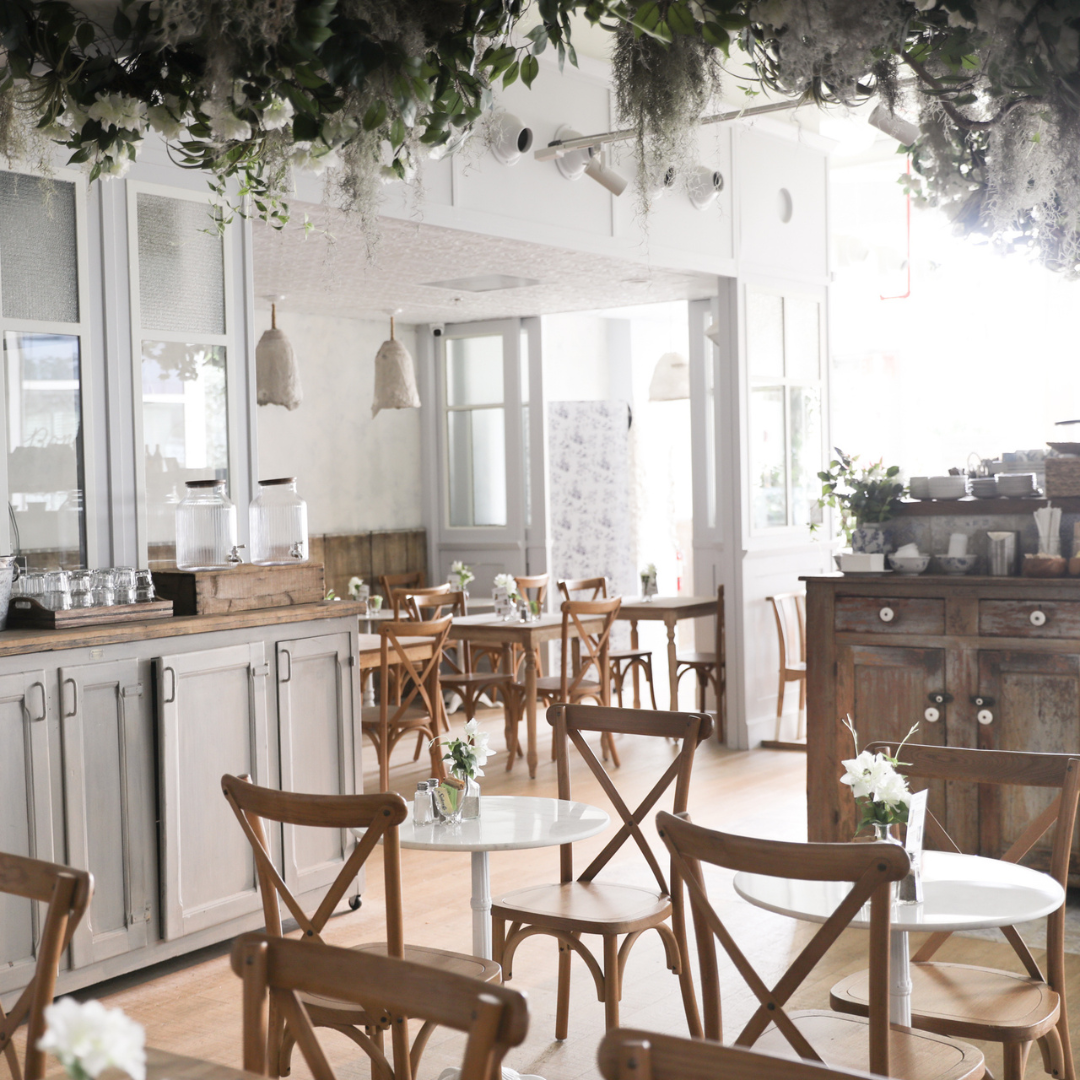 Sunlight moves through the dining room, illuminating marble tabletops, weathered wood, soft florals, and quiet corners arranged for lingering over coffee or a meal.
