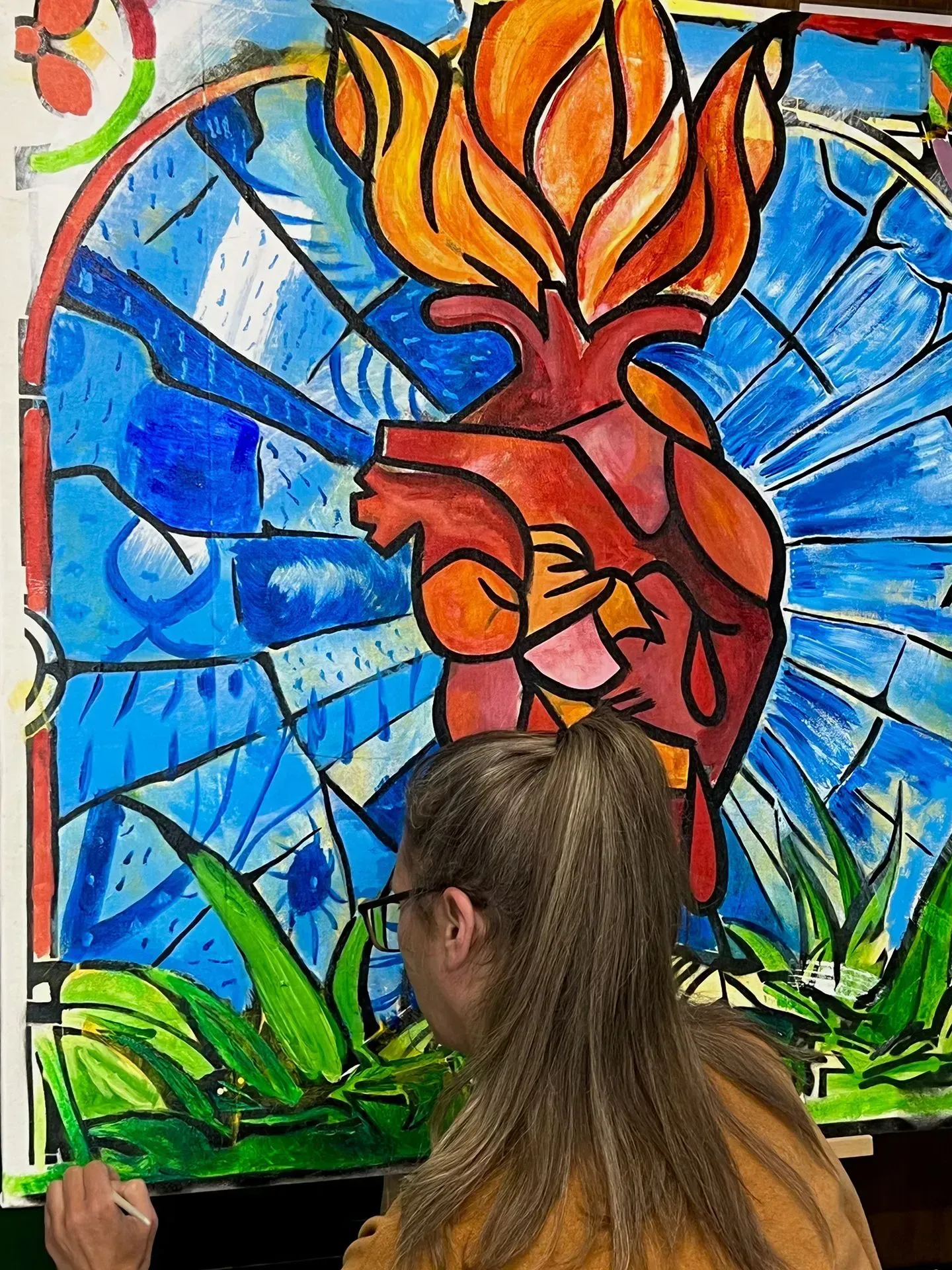Participant paints a vibrant sacred heart mural featuring bold colors, stained-glass inspiration, and symbolic artwork representing healing, creativity, hope, resilience, recovery, and personal transformation through collaborative community artistic expression.