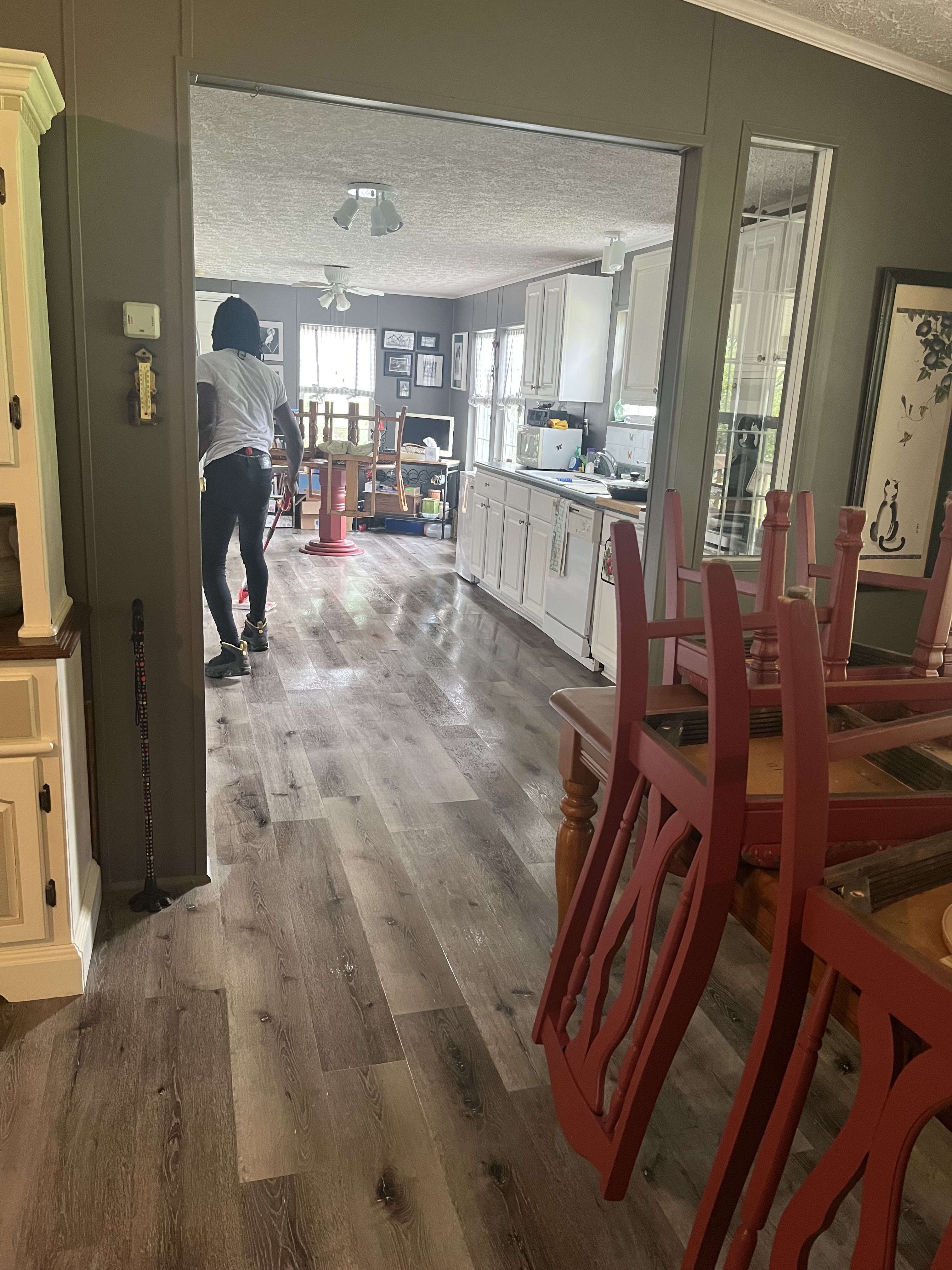 A person wearing a white t-shirt and black pants is mopping a wooden floor in a kitchen and dining area. The room is furnished with white cabinets, a stove, a sink, a dining table with red chairs, and a variety of decorative items on the walls and shelves. A thermometer is mounted on the wall near the entrance to the kitchen. Stacks of chairs are visible in the foreground.