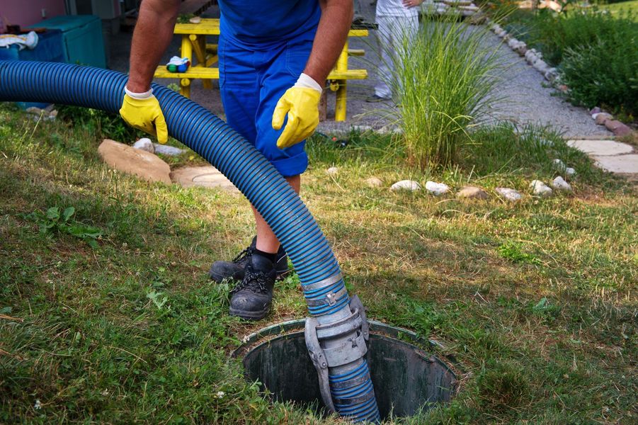 A close-up of a worker in blue overalls and yellow gloves maneuvering a thick blue suction hose into an open septic tank riser in a residential yard.
