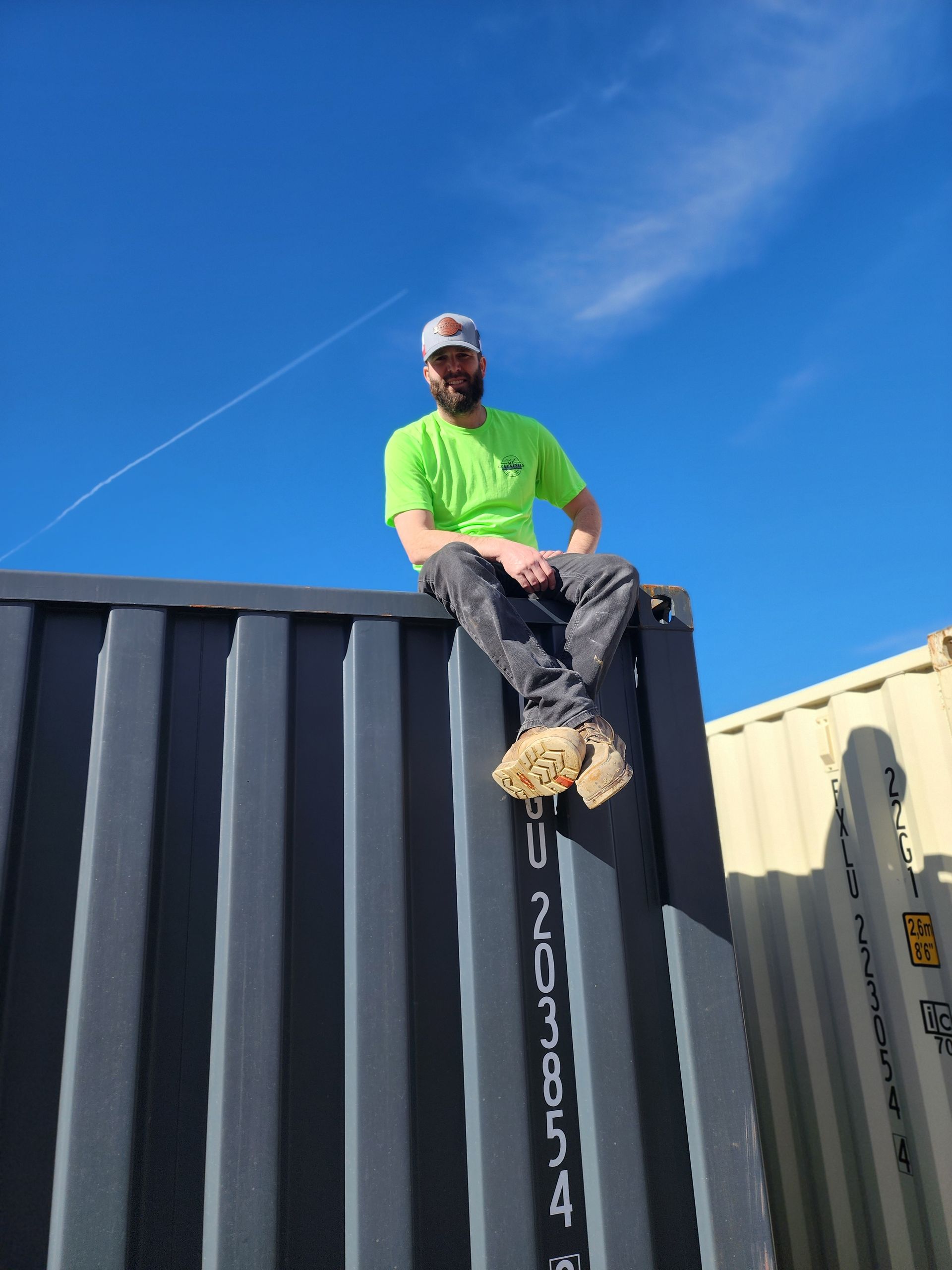 Worker sitting on top of portable storage container under clear blue sky, highlighting durability, strength, and stackable design of storage units used for transportation, storage, and construction site applications.