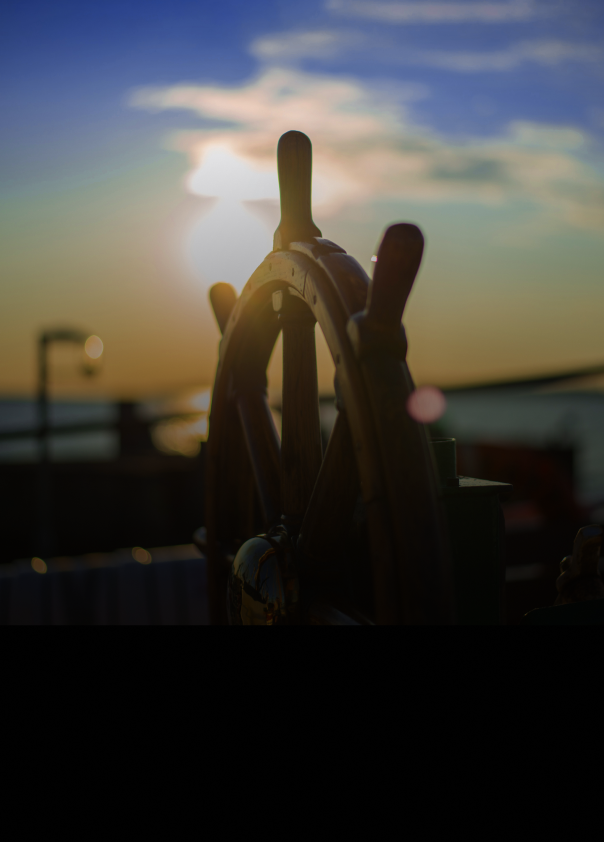 Close-up of a ship’s wooden steering wheel, with the sun setting over water in the background.