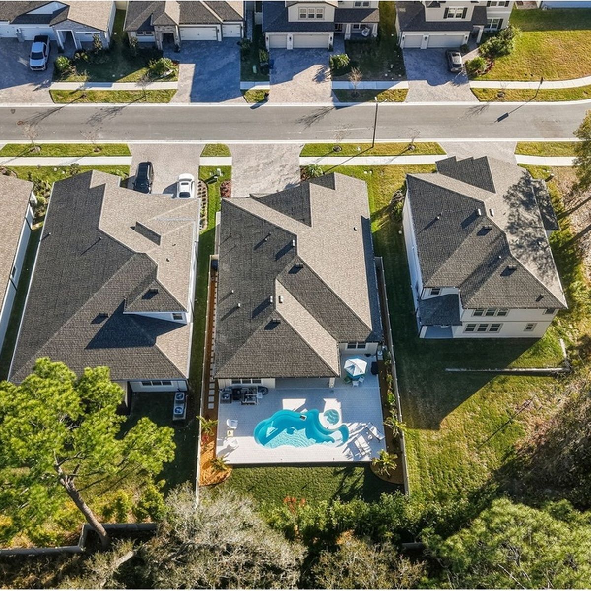 Bird's-eye view of suburban homes with dark shingle roofs, showcasing a central house featuring a custom backyard swimming pool, patio, and surrounding green lawn.