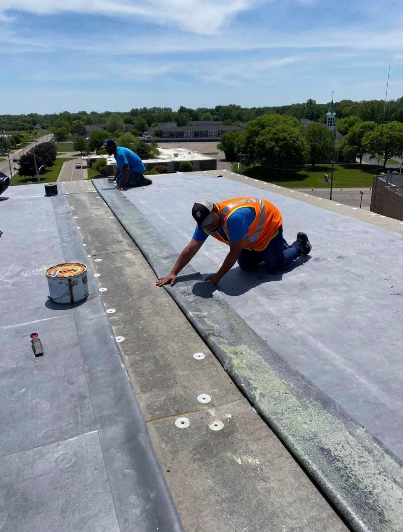 Two roofers are shown working on a large, flat roof under a bright sky. One worker in the foreground, wearing an orange safety vest, installs or seals dark roofing material.