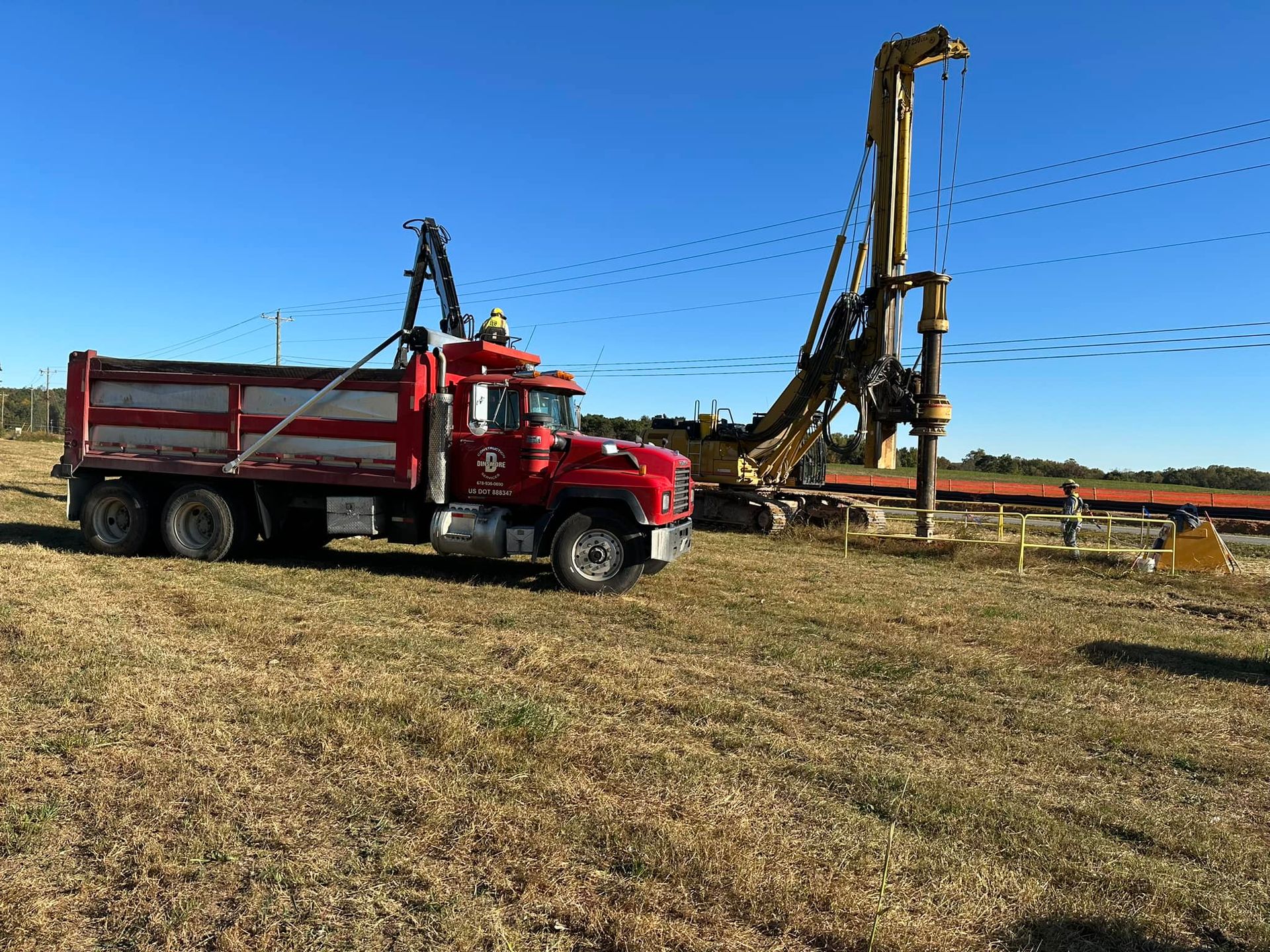A red dump truck is parked beside a large drilling rig at a construction site. Workers operate the equipment in an open field under clear blue skies.