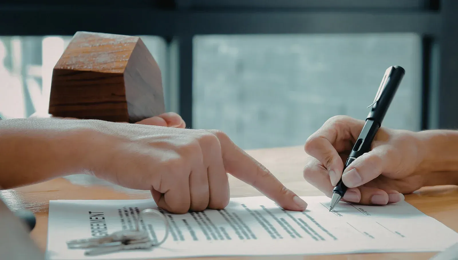 Client signing agreement while another person points at document with keys nearby, representing real estate purchase, contract review, property transfer, and final steps of secure closing transaction process.