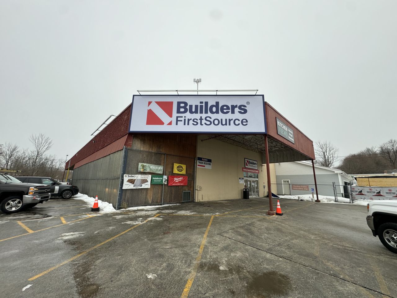 Exterior of a Builders FirstSource building materials location with large branded signage, featuring a covered front entrance, parking area, delivery access, and winter conditions outside a professional lumber and building supply facility.