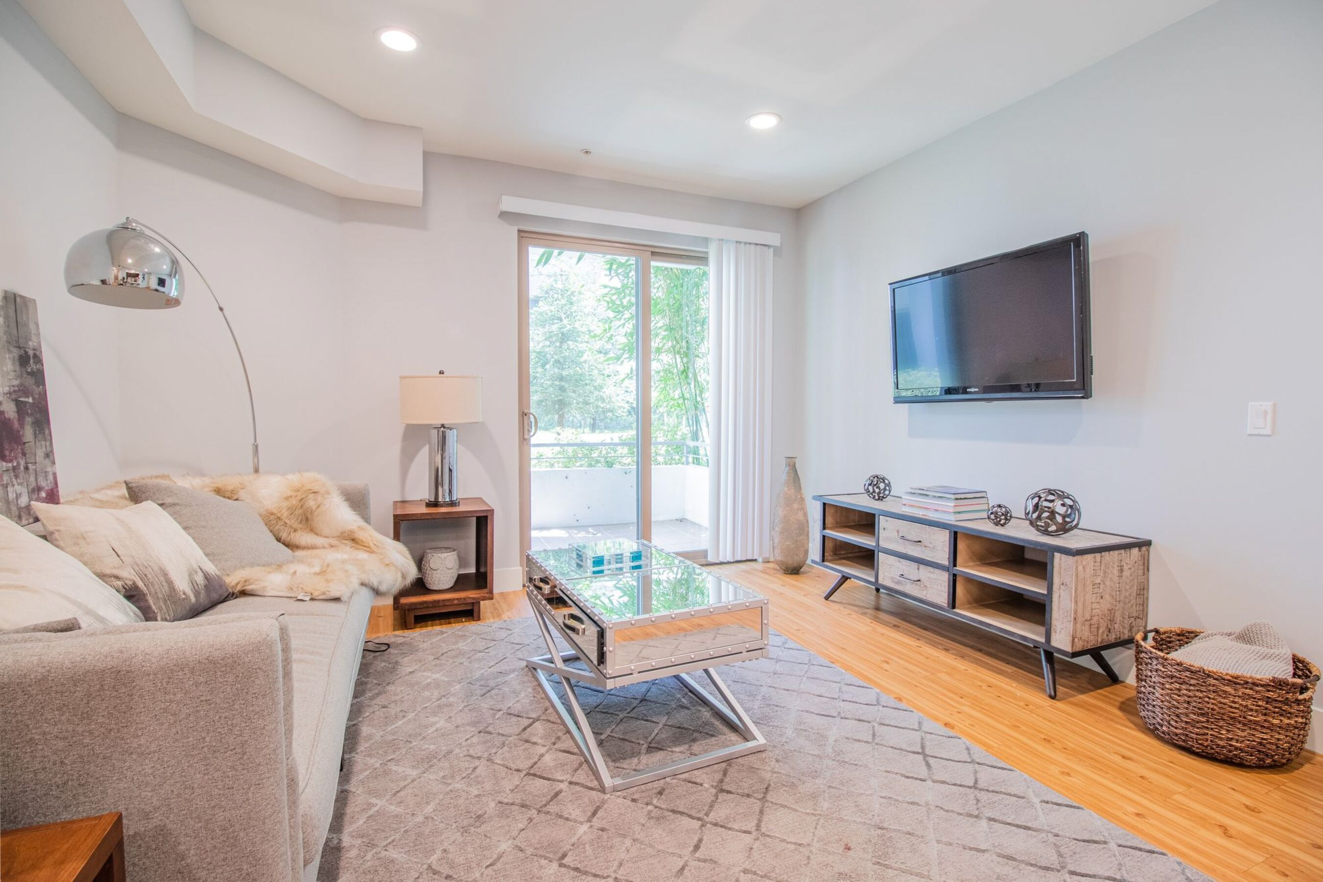 A modern living room with a grey sofa, a glass coffee table, a wooden cabinet, and a flat-screen TV mounted on the wall.