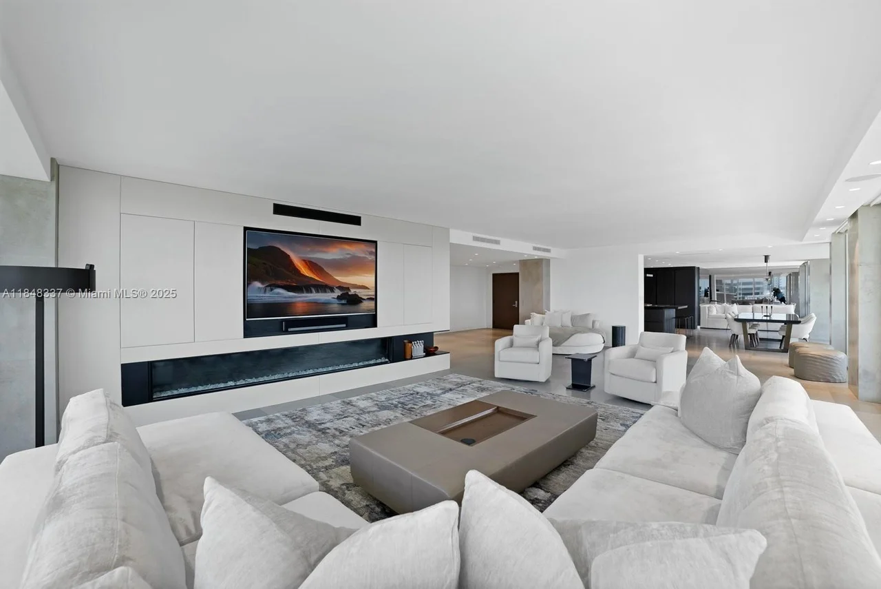 A spacious and modern living room with white sofas, a large television above a fireplace, a rectangular coffee table, and a patterned rug. In the background, there is a dining area and kitchen visible. The room is decorated in a minimalist style with neutral colors.