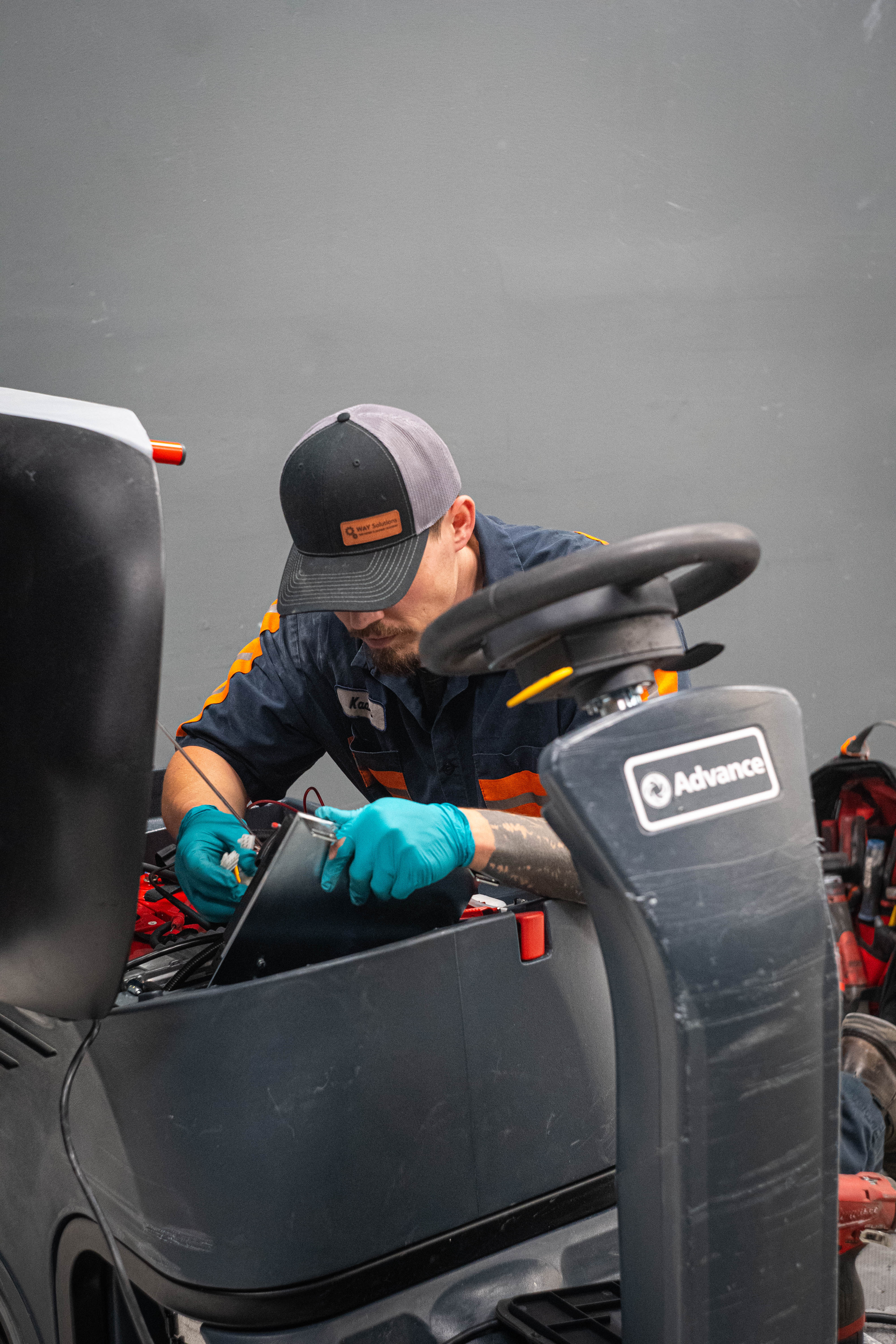 Equipment Model: Advance Ride-On Commercial Floor Scrubber
Action: Internal control console and electrical wiring harness repair
Personnel: Lead technician Kaden wearing protective teal nitrile gloves and a branded WaySolutions cap
Service Focus: Complex electrical troubleshooting and control panel maintenance