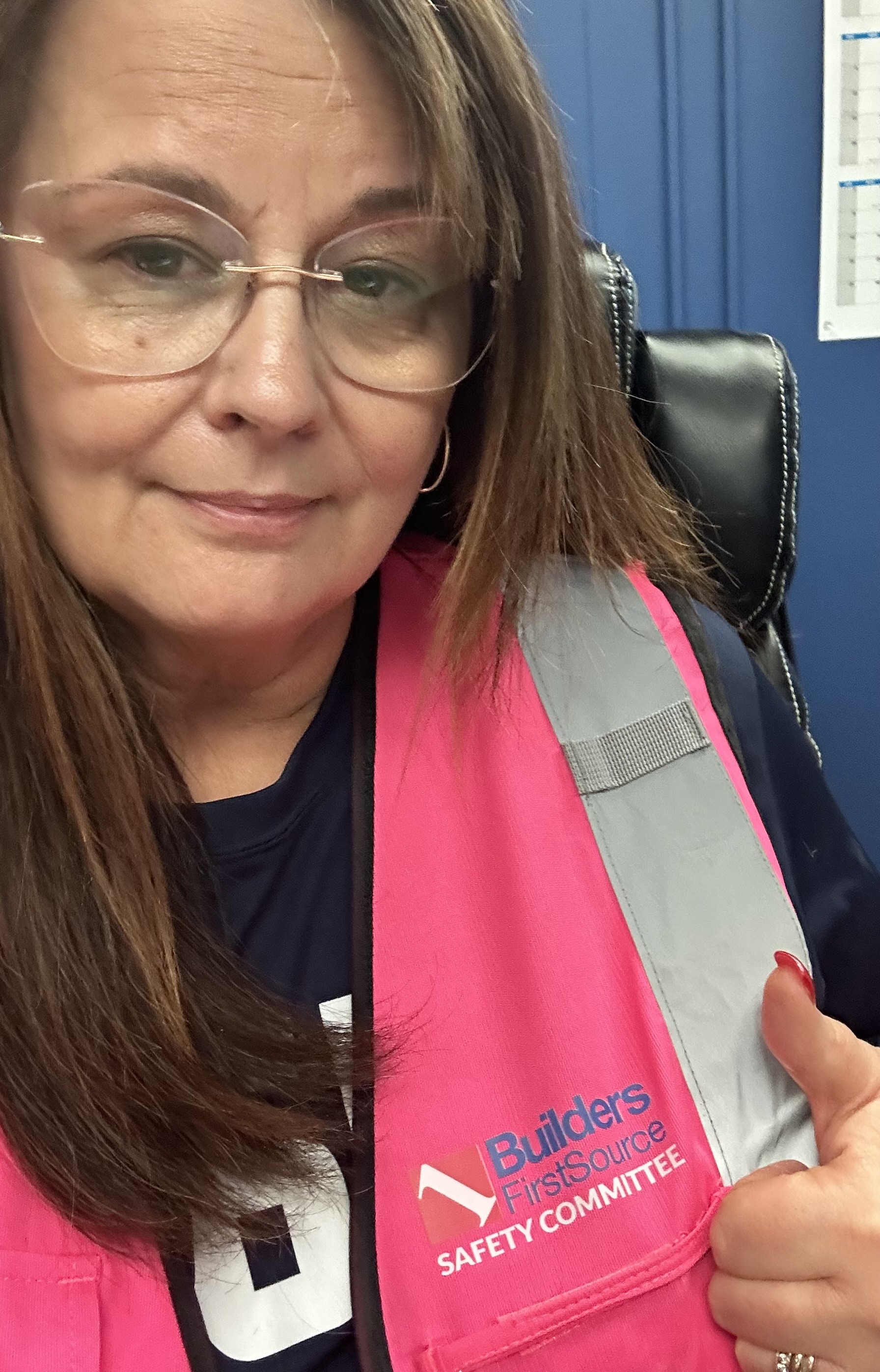 A close-up selfie of a woman wearing glasses and a bright pink safety vest with the "Builders FirstSource Safety Committee" logo on the front. She has long brown hair and is smiling slightly, giving a thumbs-up with her right hand. The background shows a blue office wall and part of a desk, suggesting she is in a workplace environment. The overall tone of the image reflects confidence and positivity regarding her role in the safety committee.