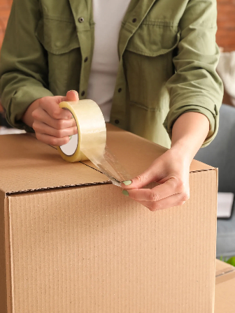 Close-up shot of hands using clear packing tape to securely seal the top seam of a corrugated cardboard moving box.
