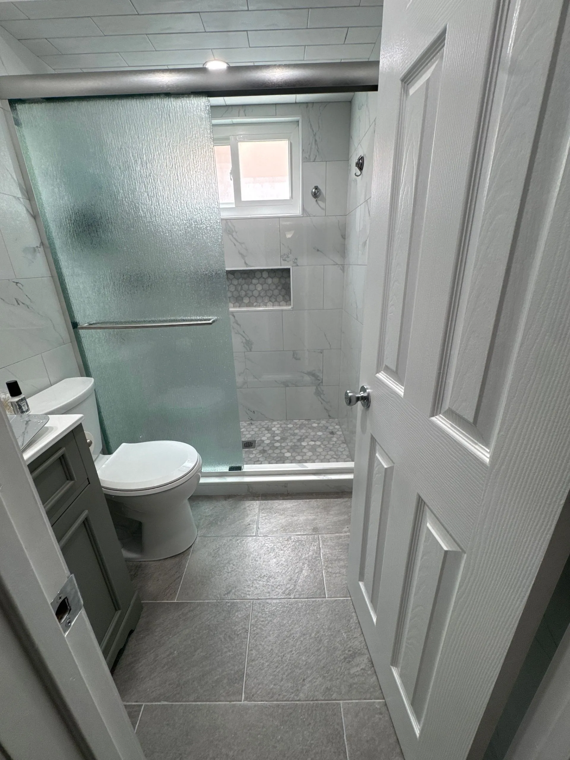 Renovated bathroom showcasing a walk-in shower with textured glass sliding door, tile niche, and modern finishes. The remodel enhances comfort, accessibility, and visual appeal throughout.